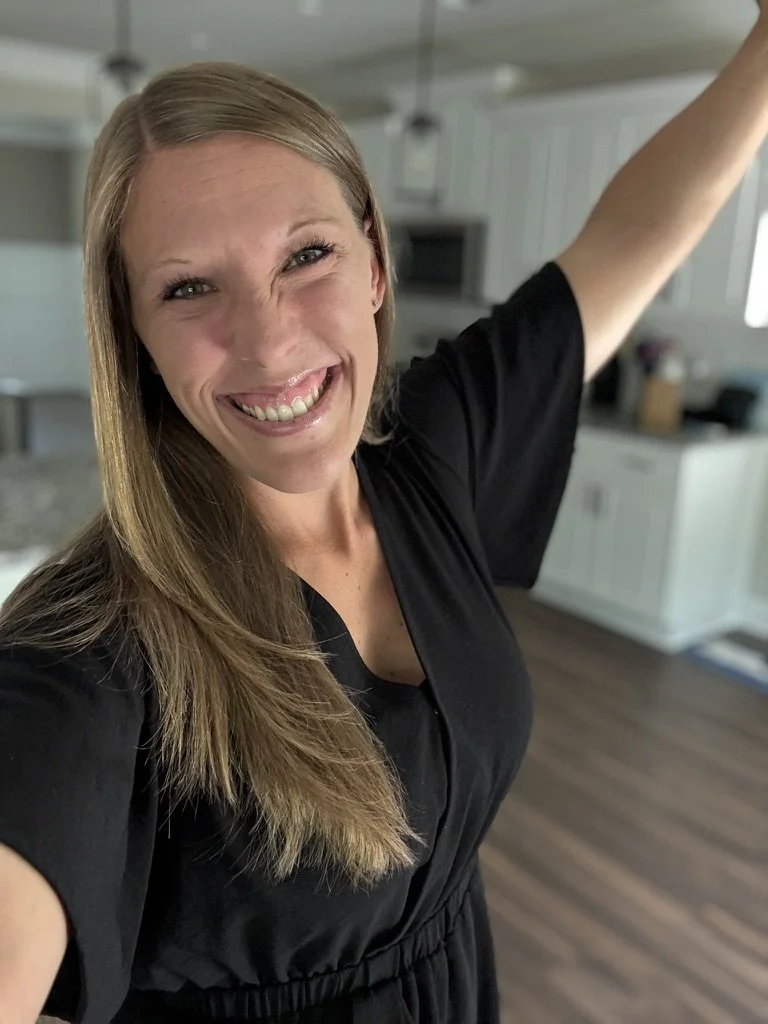 A woman with long blonde hair smiling and raising her arm in a kitchen.