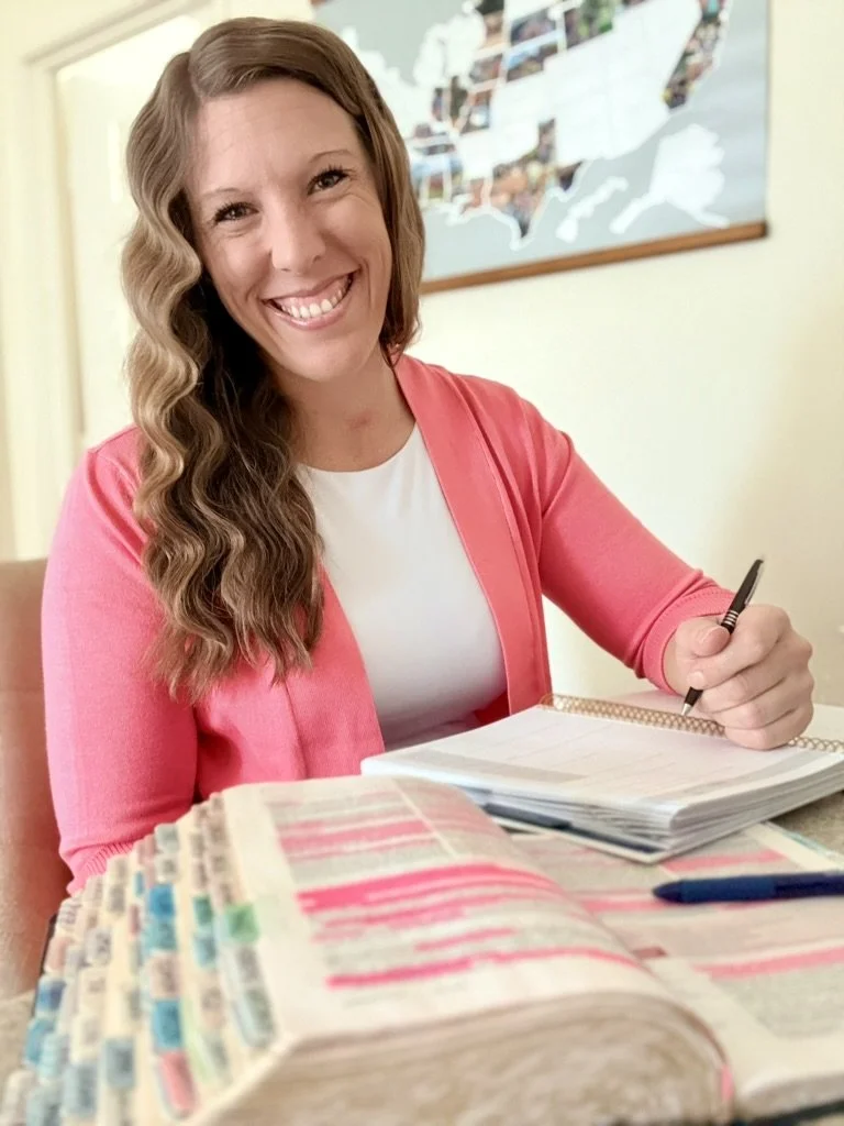 A woman with long, wavy brown hair and a bright smile is sitting at a desk with a large open book and a notepad. She is wearing a white top and a pink cardigan. Behind her, there is a map of the United States on the wall.