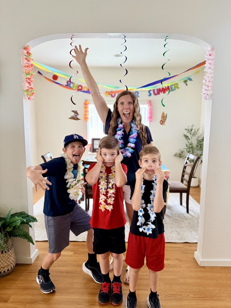 A woman and three boys celebrating a summer party with leis, colorful streamers, and a 'SUMMER' banner in the background, in a decorated living room.