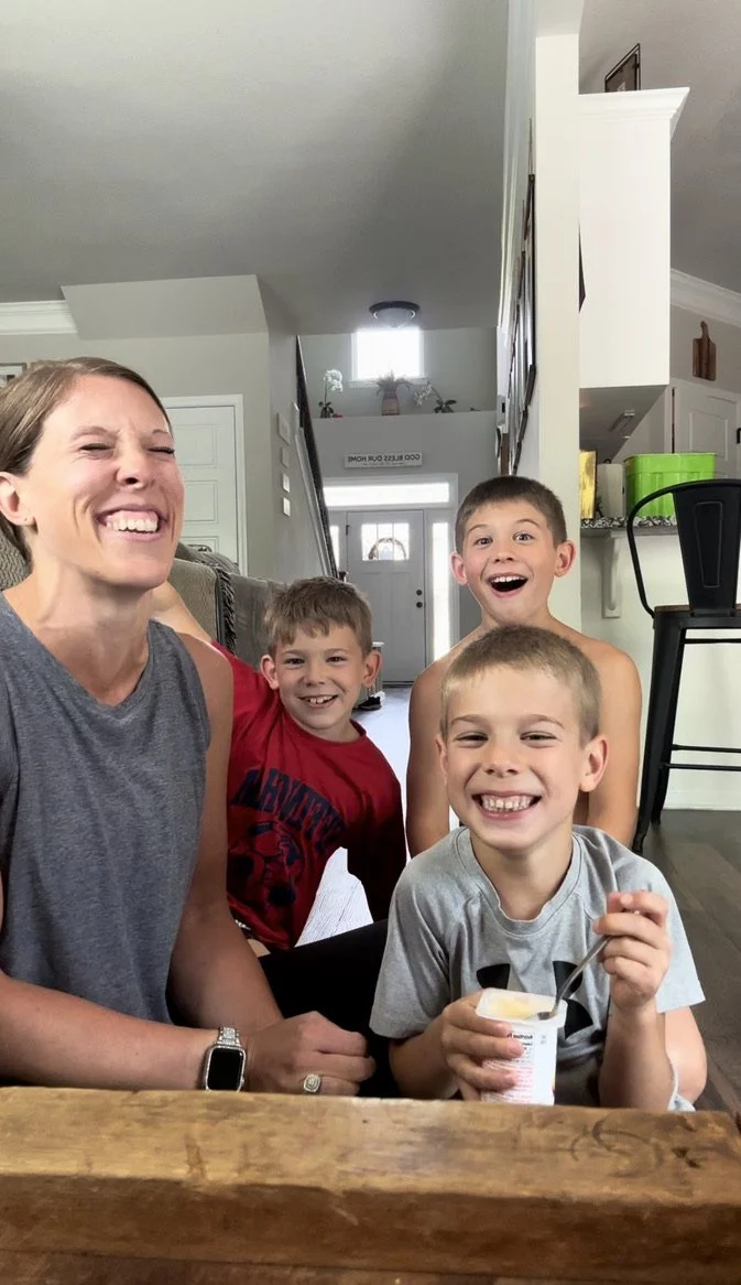 A woman and three young boys smiling and laughing in a cozy home kitchen or dining area. The woman has short hair and is wearing a sleeveless grey top, while the boys have short hair and are wearing casual grey and red T-shirts. One boy is holding a cup of yogurt with a spoon.