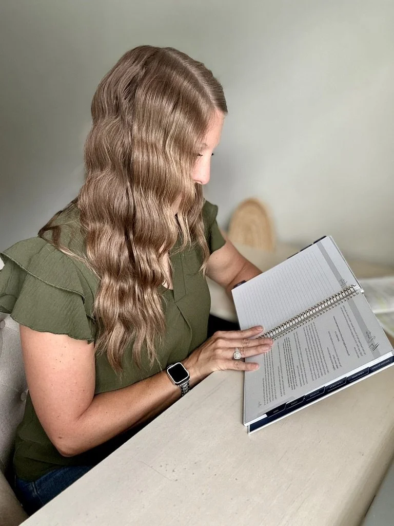 Woman with long wavy hair reading a spiral-bound book at a table.