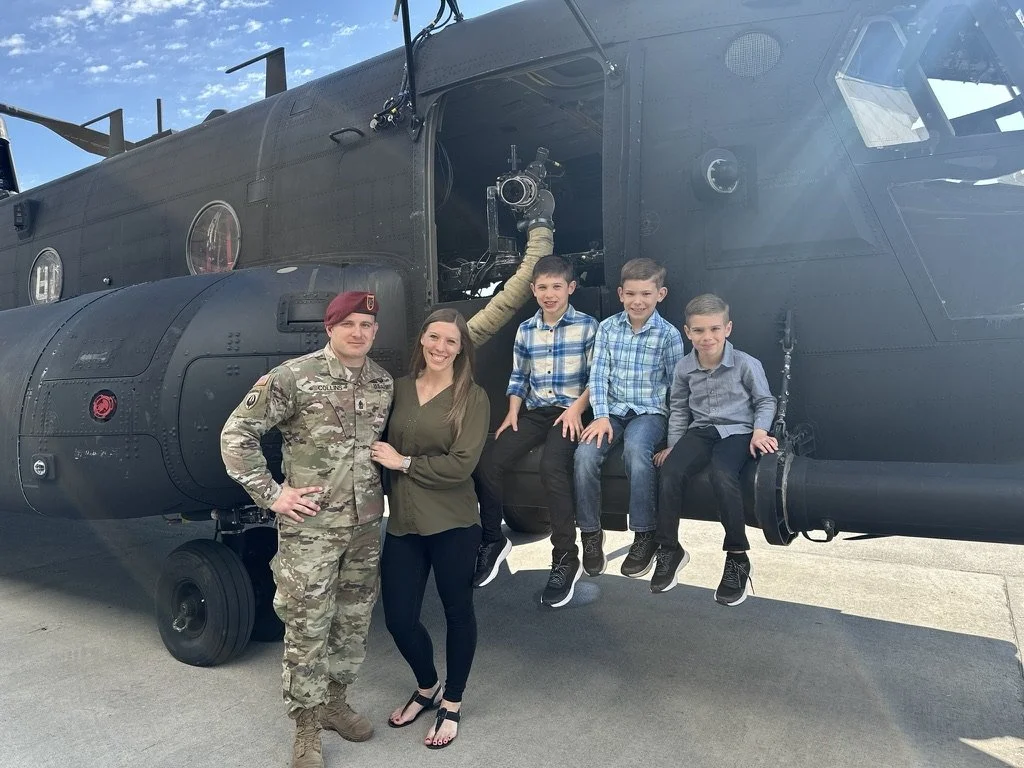 A soldier and a woman are standing next to a military helicopter on a tarmac, with three children sitting on the helicopter's side.
