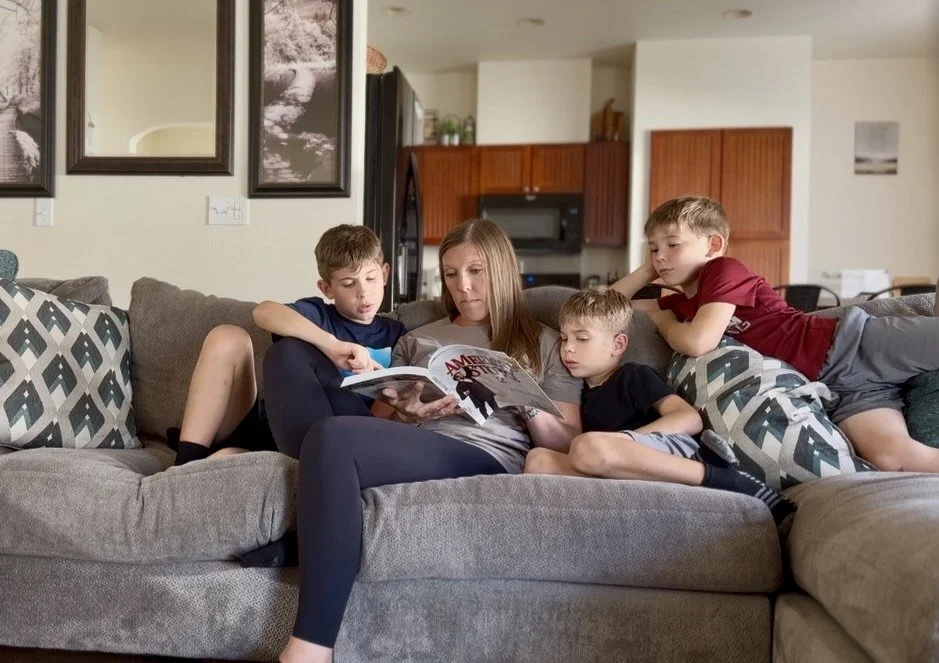 A woman reading a magazine on a gray couch with three young boys gathered around her, in a living room with art on the wall and a kitchen in the background.