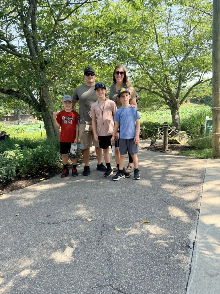 Family of five standing outdoors on a shaded path with trees and greenery in the background.
