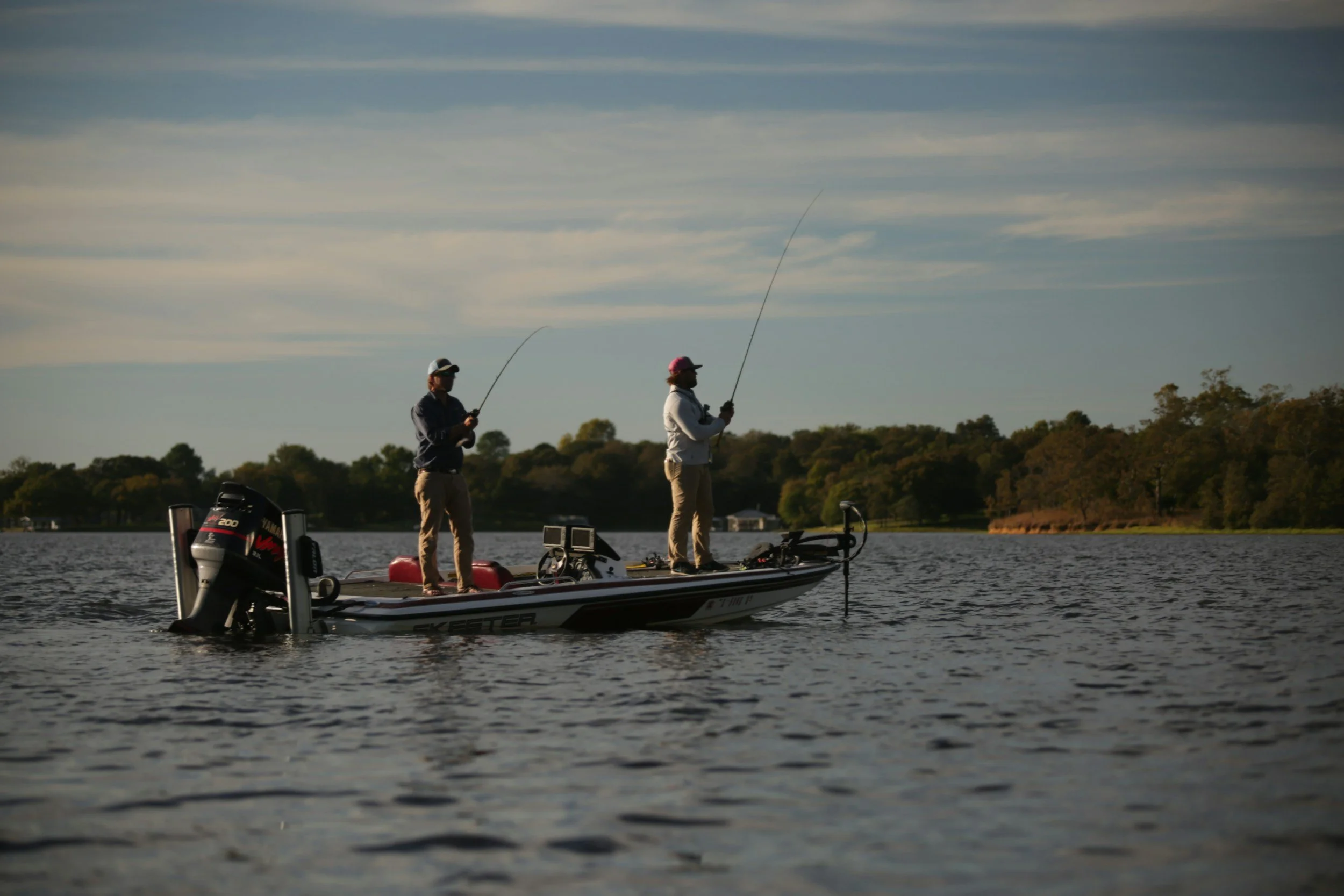 Two men fishing from a boat on a lake in the late afternoon, with trees and houses in the background.