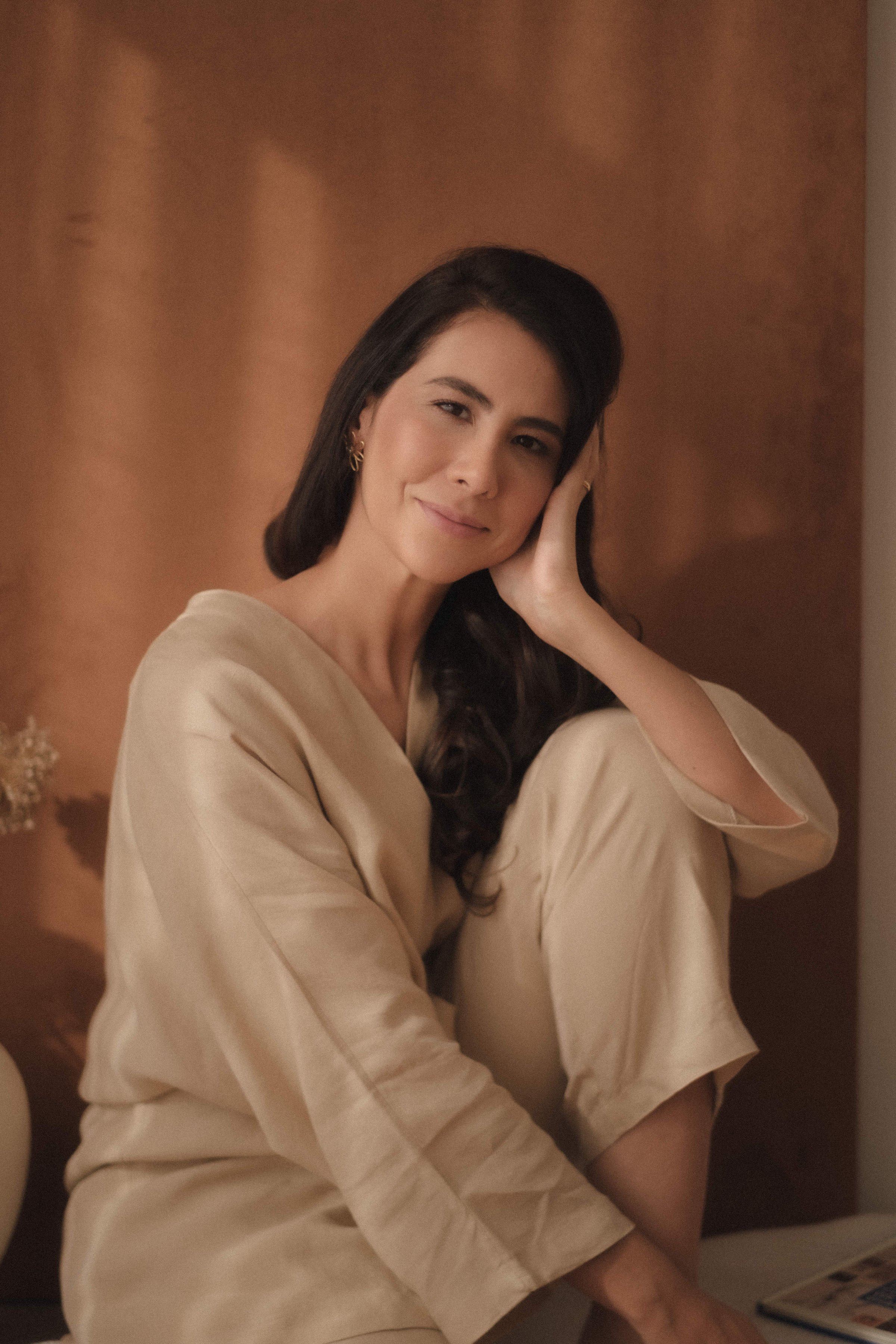 A woman sitting in a relaxed pose, smiling gently with one hand resting on her cheek. She has long dark hair and is wearing earrings and a beige, loose-fitting outfit. The background features a brown wall with minimal decoration.