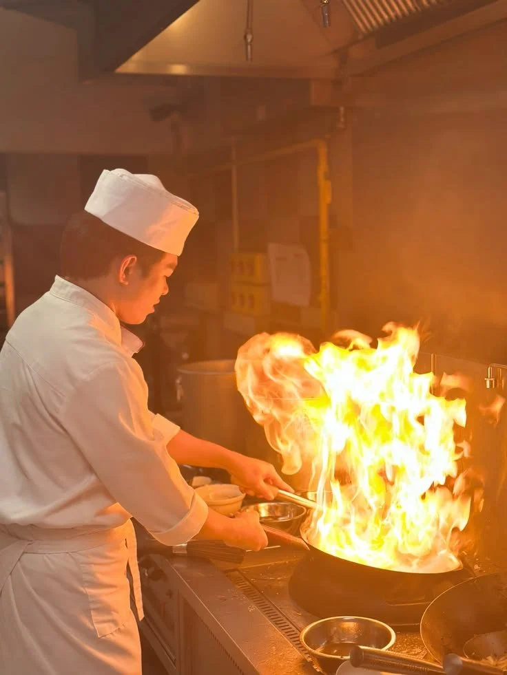 Chef in white uniform and hat cooking with a large flame erupting from a wok.