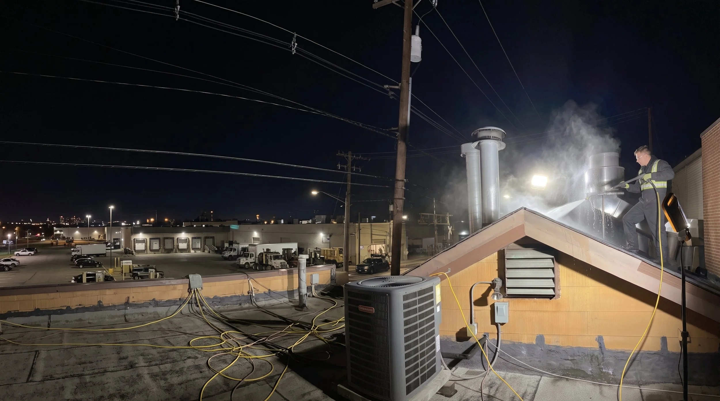 A man washing the roof of a building at night with a pressure washer, emitting steam or water vapor, in an industrial or commercial area with parked trucks and warehouses in the background.