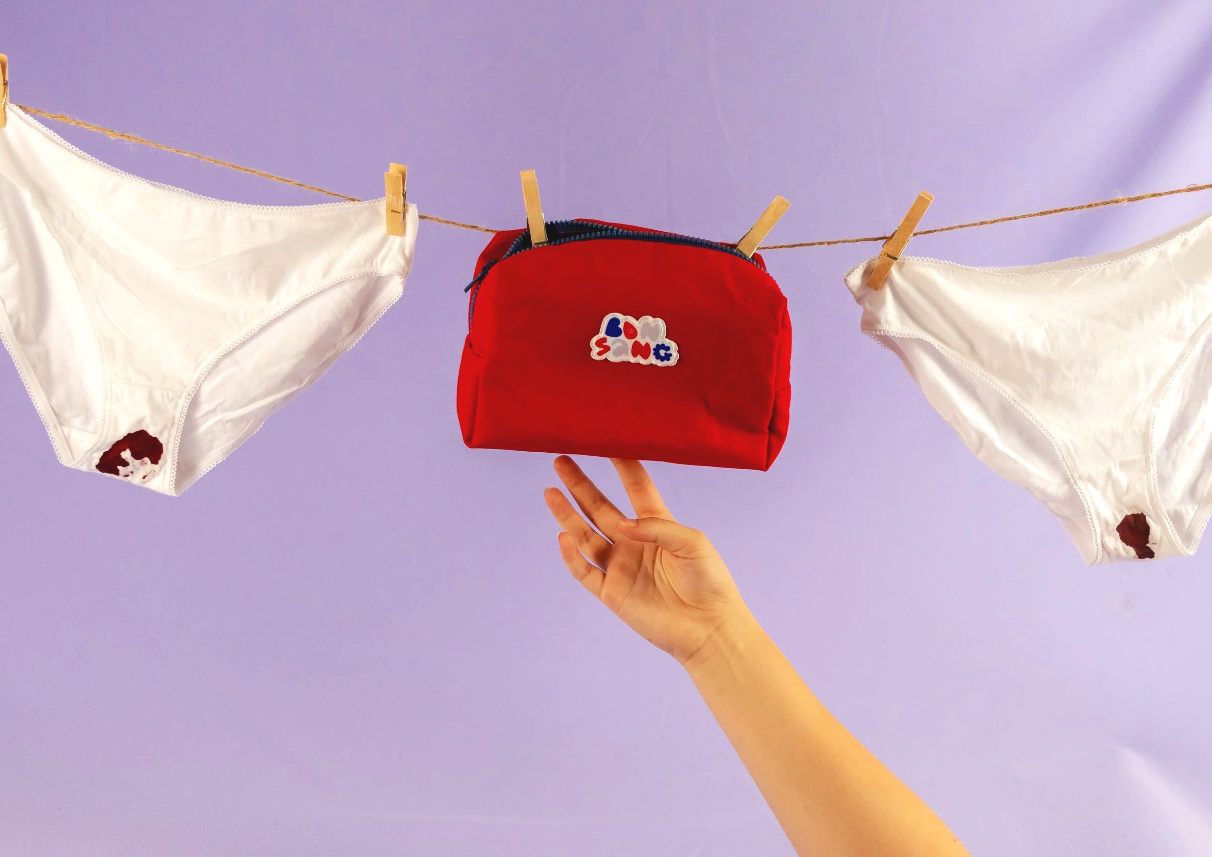 L'image montre une corde à linge avec deux vêtements blancs suspendus à l'aide d'épingles en bois et un sac rouge avec un patch imprimé Love et Peace. La main d'une personne tient le sac. Le ciel est violet clair.