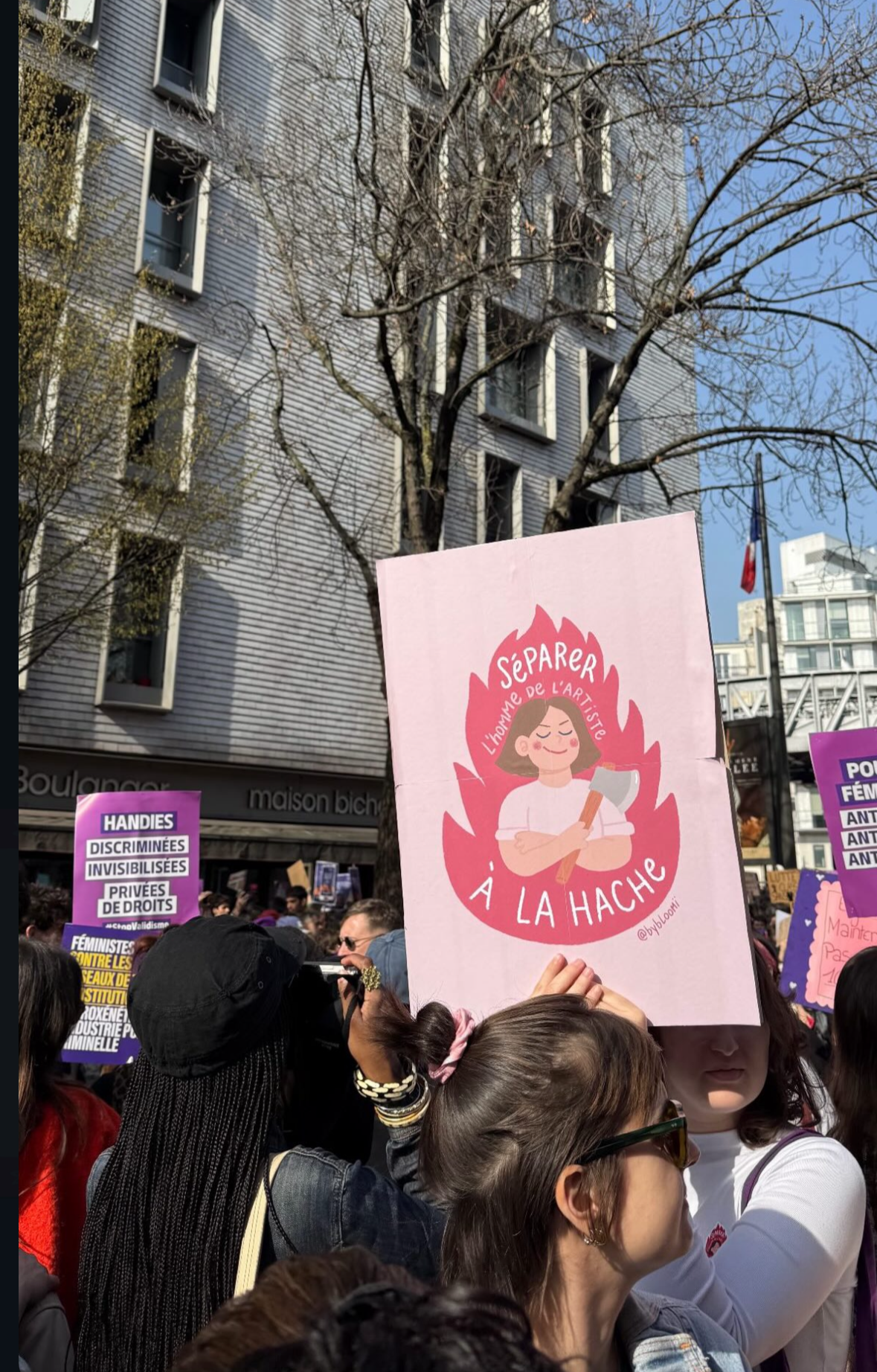 Une manifestation avec des pancartes, dont une tête une femme souriante avec une hache dans un feu, portant le texte "Séparer l'homme de l'artiste à la hache", lors d'une journée ensoleillée dans une ville. 
