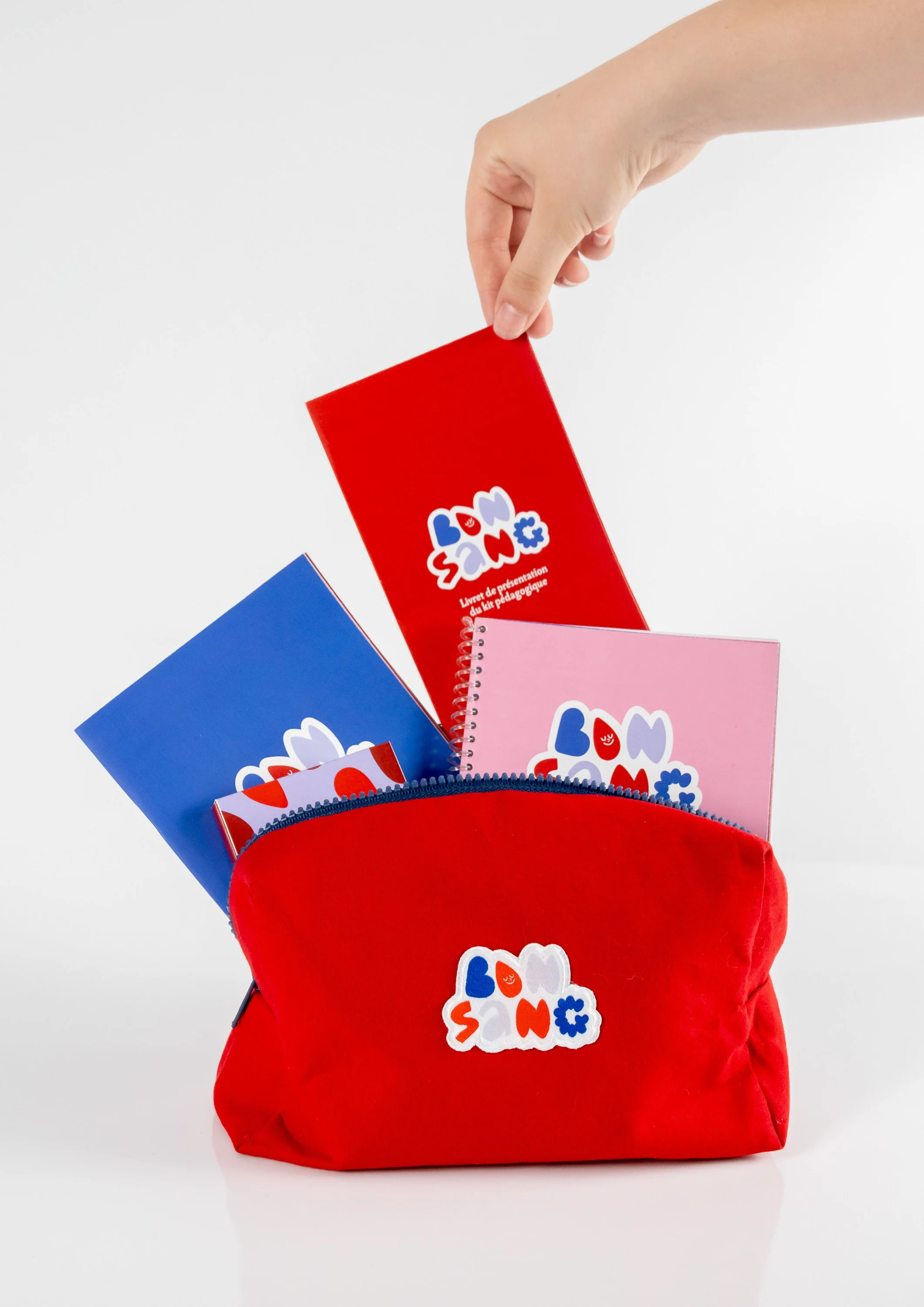 Une main tenant une bannière rouge avec un logo coloré "SANG" et trois cahiers de couleurs bleu, rose et blanc dans une poche rouge