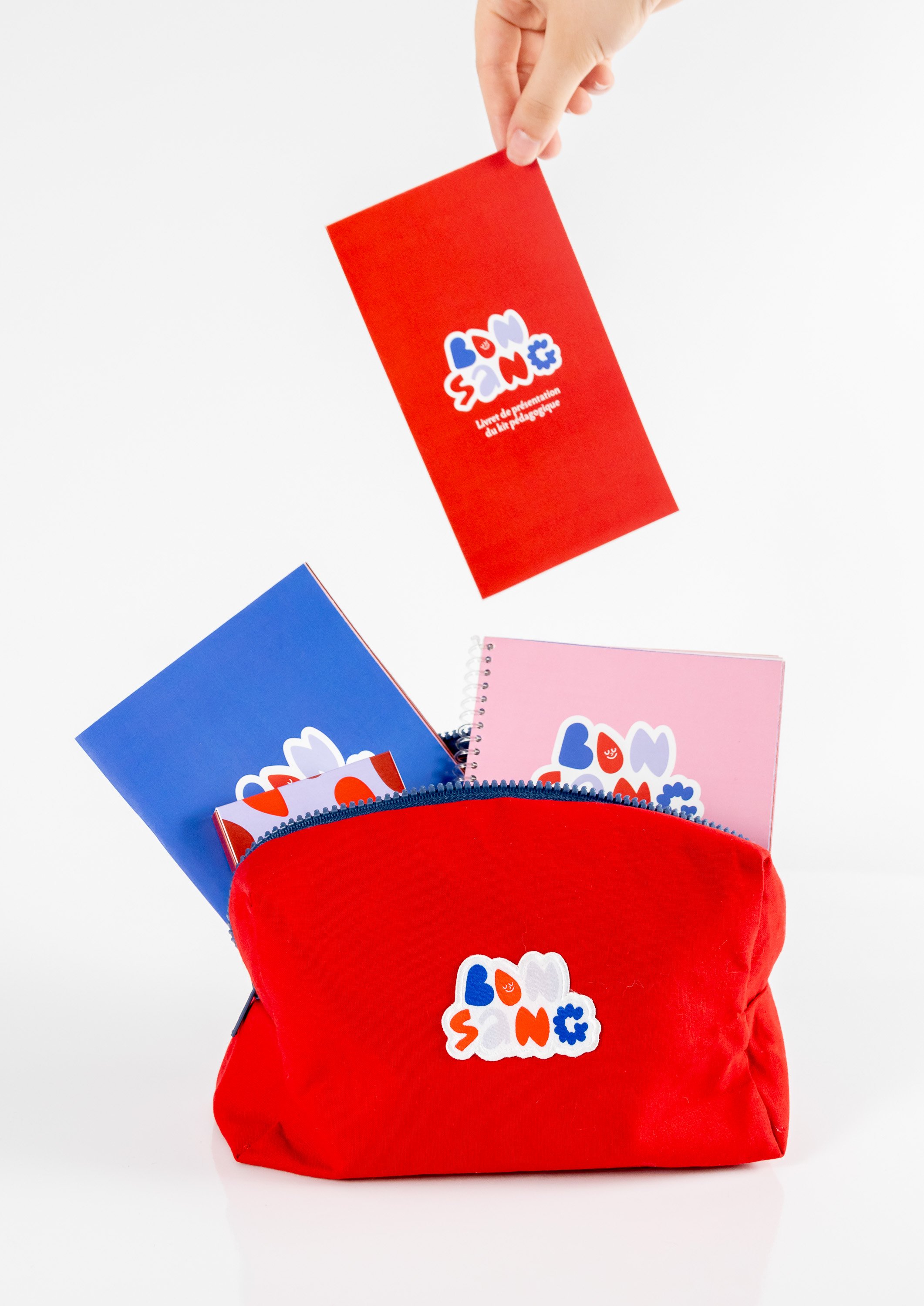 Une pochette rouge contenant plusieurs cahiers colorés (bleu, rose) avec des dessins de nuages et des lettres, le tout placé dans un sac rouge.