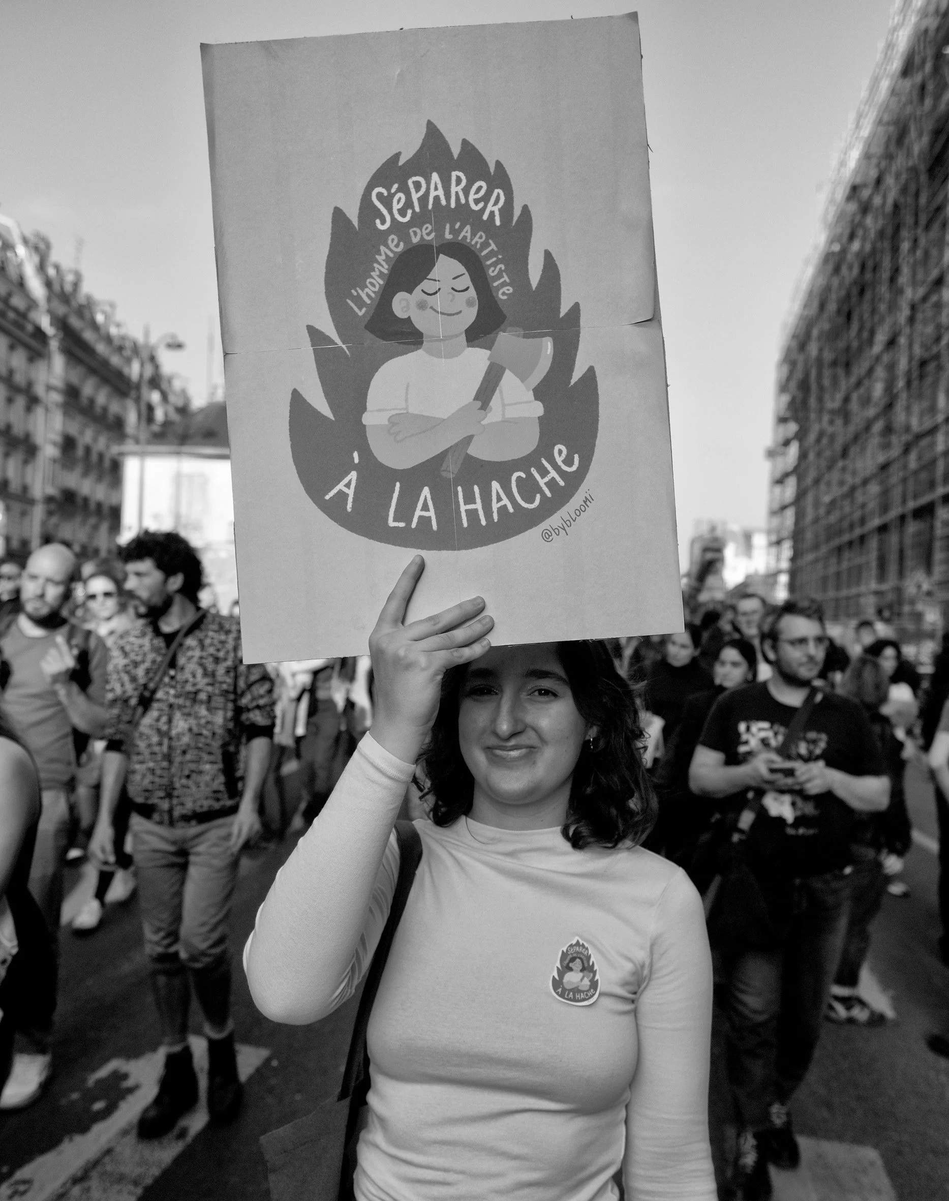 Une femme tient une affiche représentant une caricature d'une femme avec un torche, entourée d'une flamme, avec le texte "Separer l'homme de l'artiste à la hache". Elle porte un t-shirt blanc avec un badge".