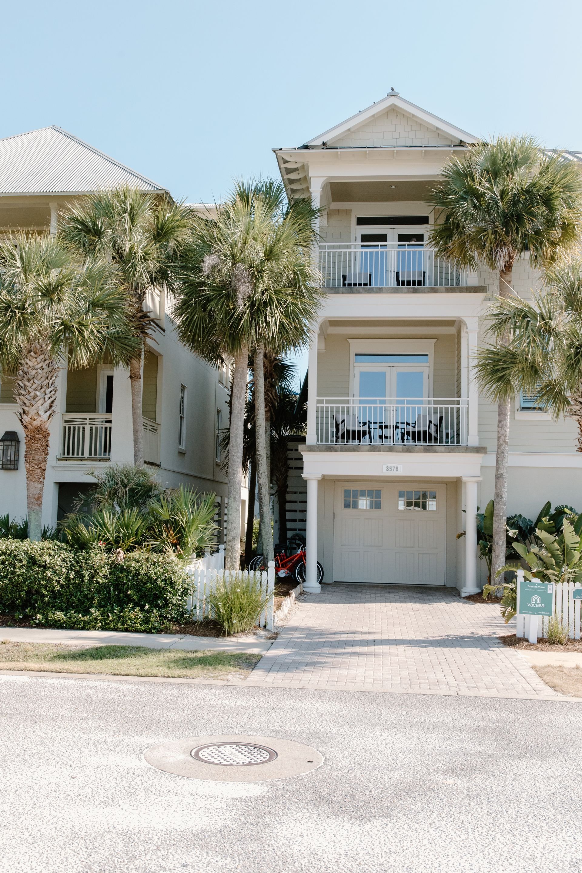 A light-colored beach house with two balconies and white railing, surrounded by palm trees, with a driveway and a garage, on a sunny day with clear blue sky.