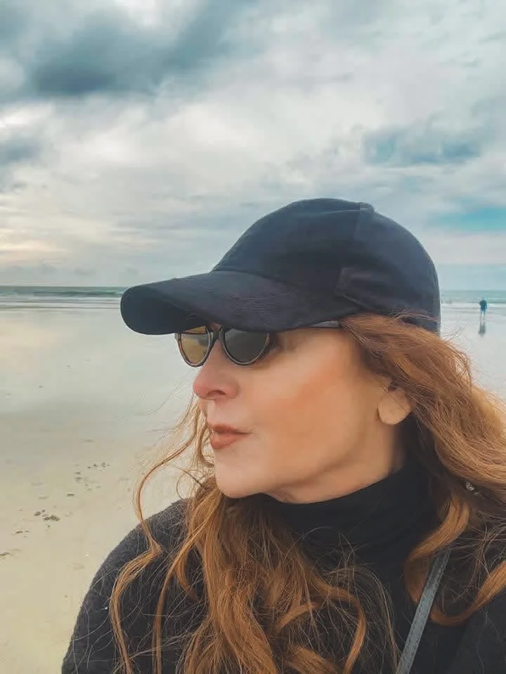Woman with auburn hair wearing black cap and sunglasses on a beach with cloudy sky.