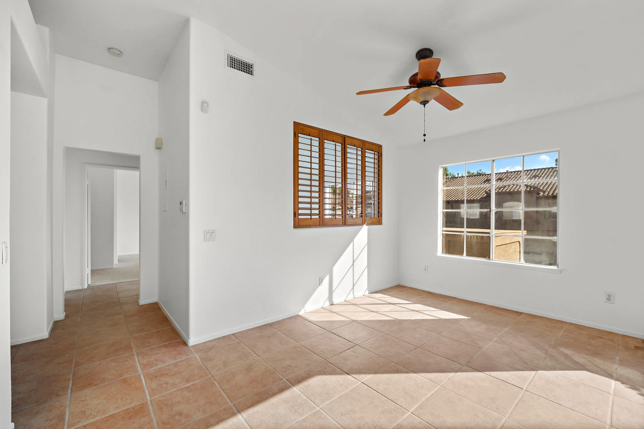 Empty living room with white walls, a ceiling fan, large windows, and tan tiled flooring.