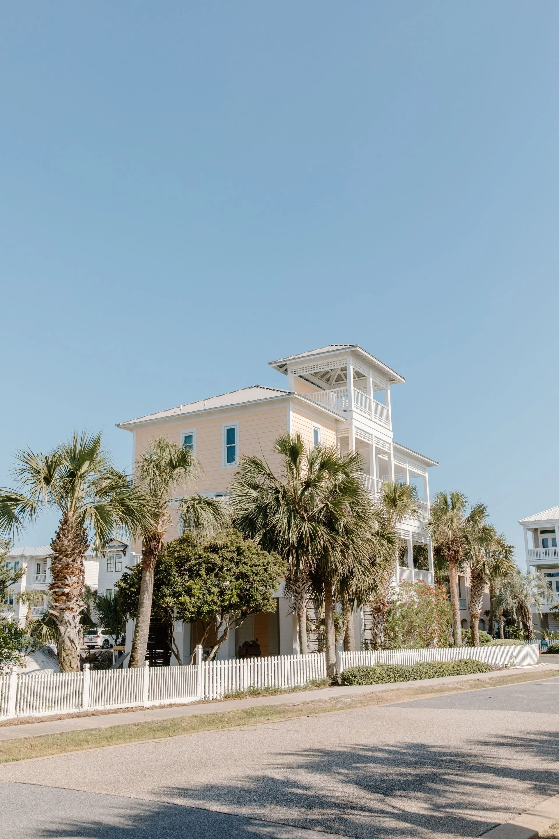 A beach house with light peach walls and white trim, surrounded by palm trees, and a white picket fence at the sidewalk, under a clear blue sky.