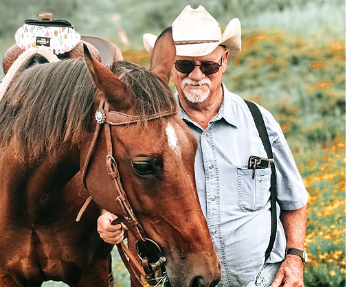 An older man wearing a cowboy hat and sunglasses stands next to a brown horse with a white stripe on its face, in a field with yellow flowers.