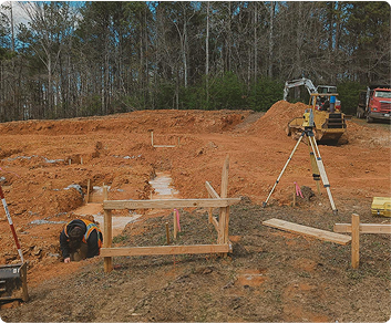 Construction site with workers, surveying equipment, and earth-moving machinery on a dirt lot, with trees in the background.