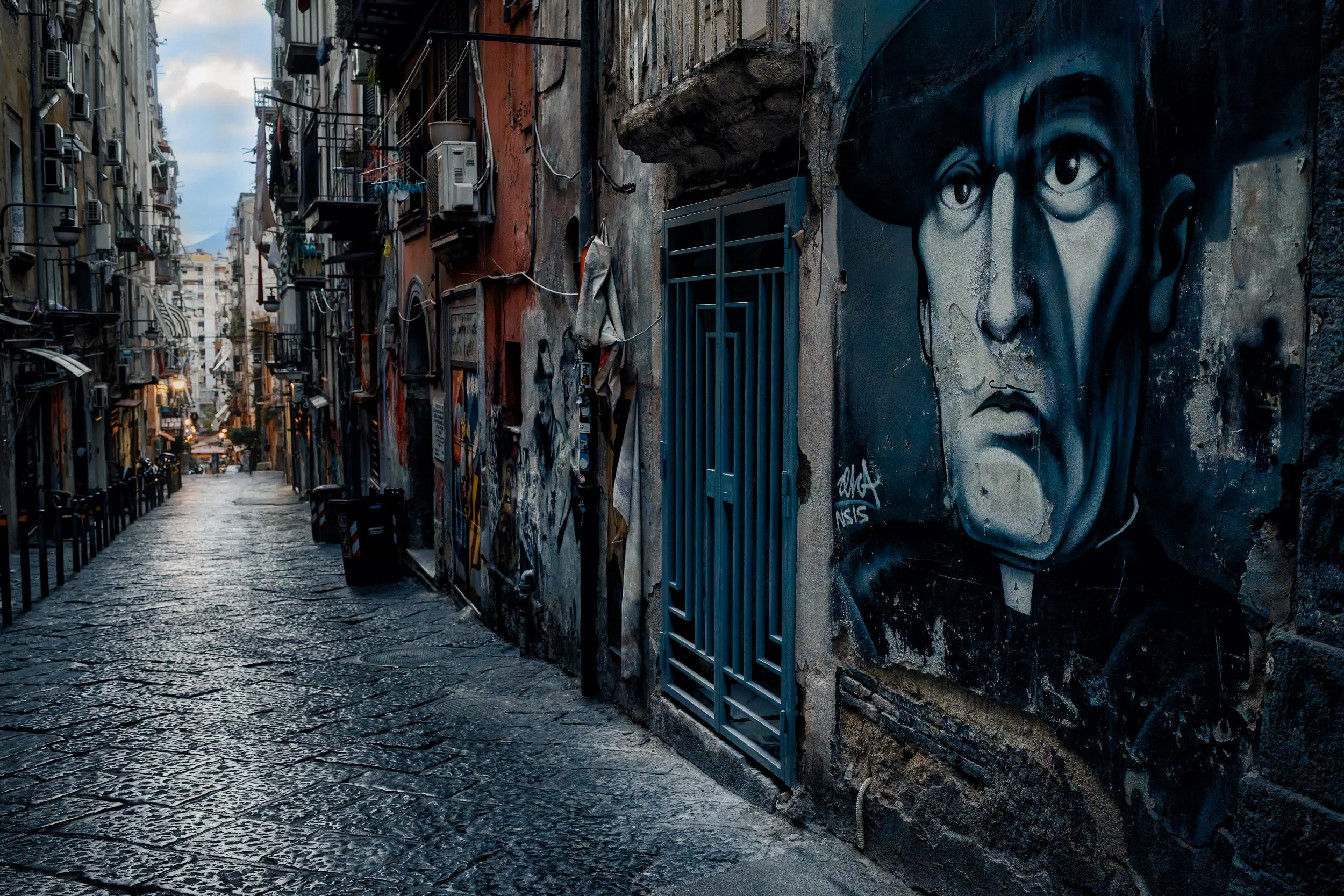 Una strada di ciottoli con edifici vecchi e affrescati. Sulle pareti ci sono graffiti, tra cui un grande murale di un volto di donna con capelli neri e occhi espressivi.