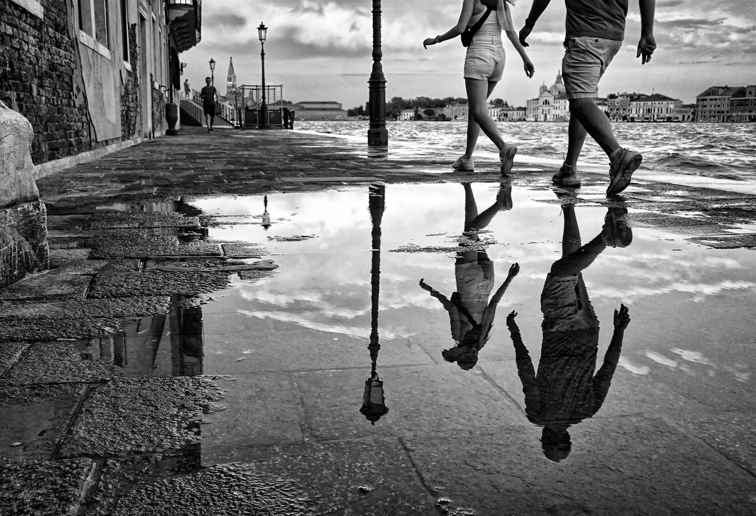 Due persone camminano lungo un molo sopra una pozzanghera che riflette il cielo e i palazzi sullo sfondo, con lampioni lungo la strada e un edificio con una torre in lontananza.