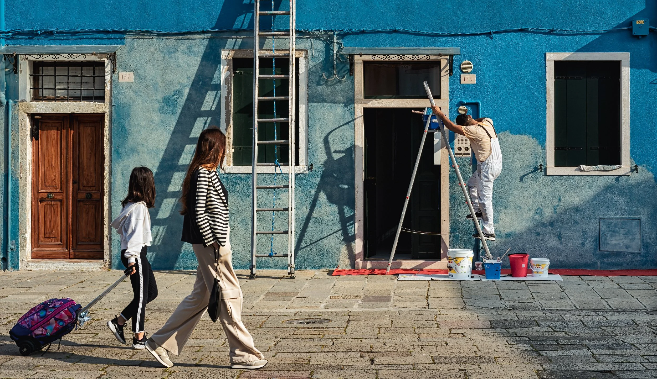 A person paints a blue façade while two people stand nearby, with ladders, windows and painting materials arranged along the pavement.
