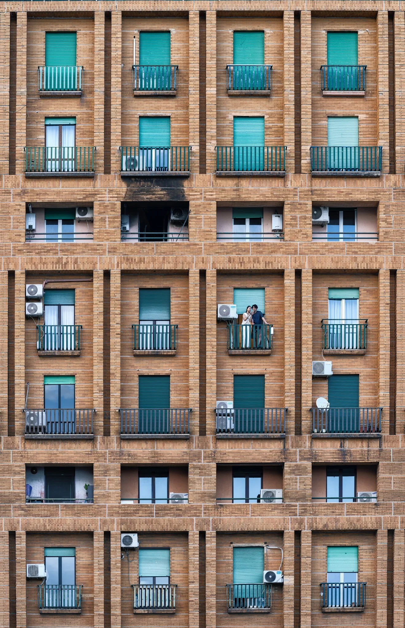 A residential façade divided into repeating balconies and windows, with two people standing neara dark stain on the building.