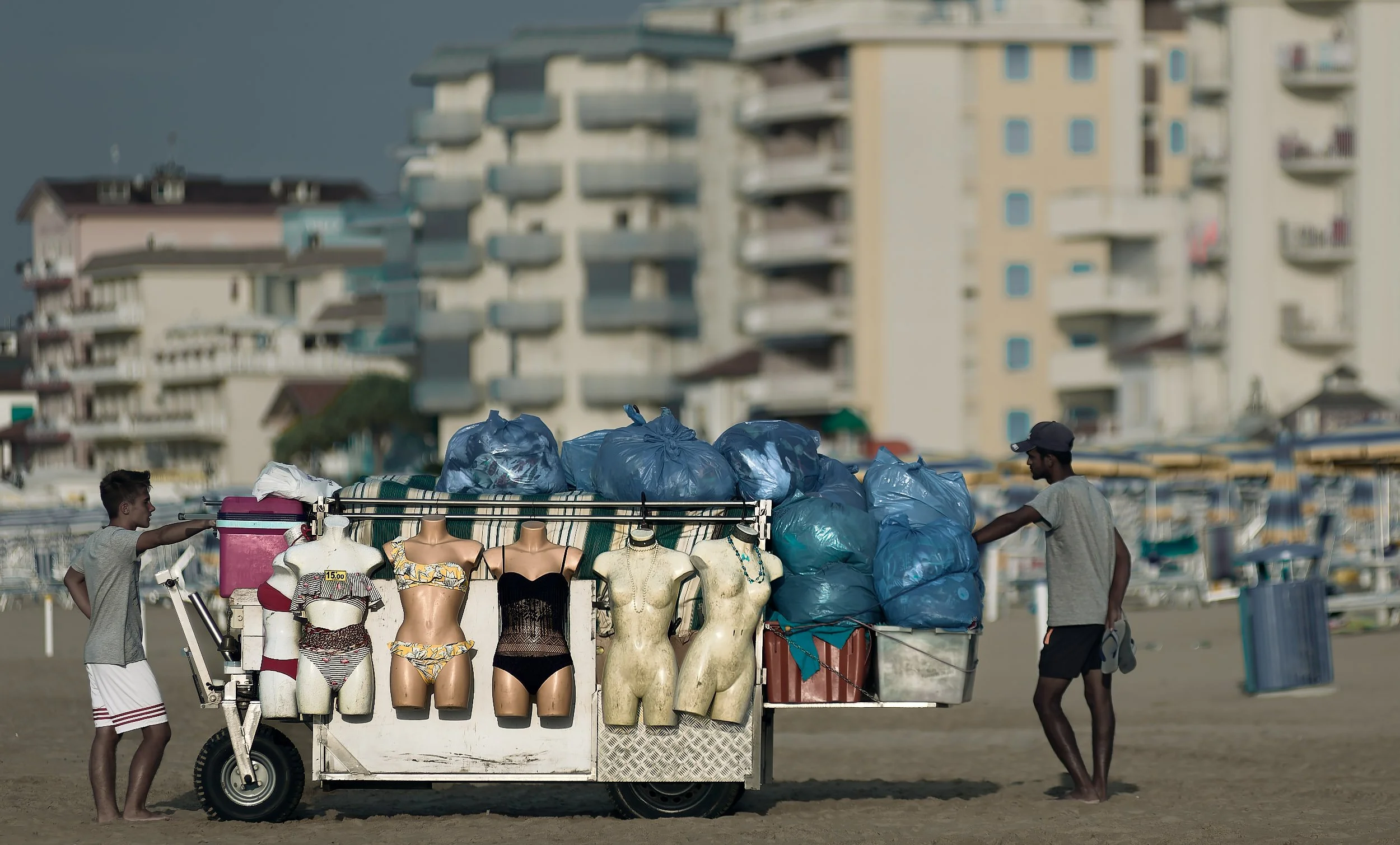 Furgoncino con manichini che indossano costumi da bagno e buste di plastica blu sopra, due uomini che si tengono per mano, sullo sfondo edifici residenziali.