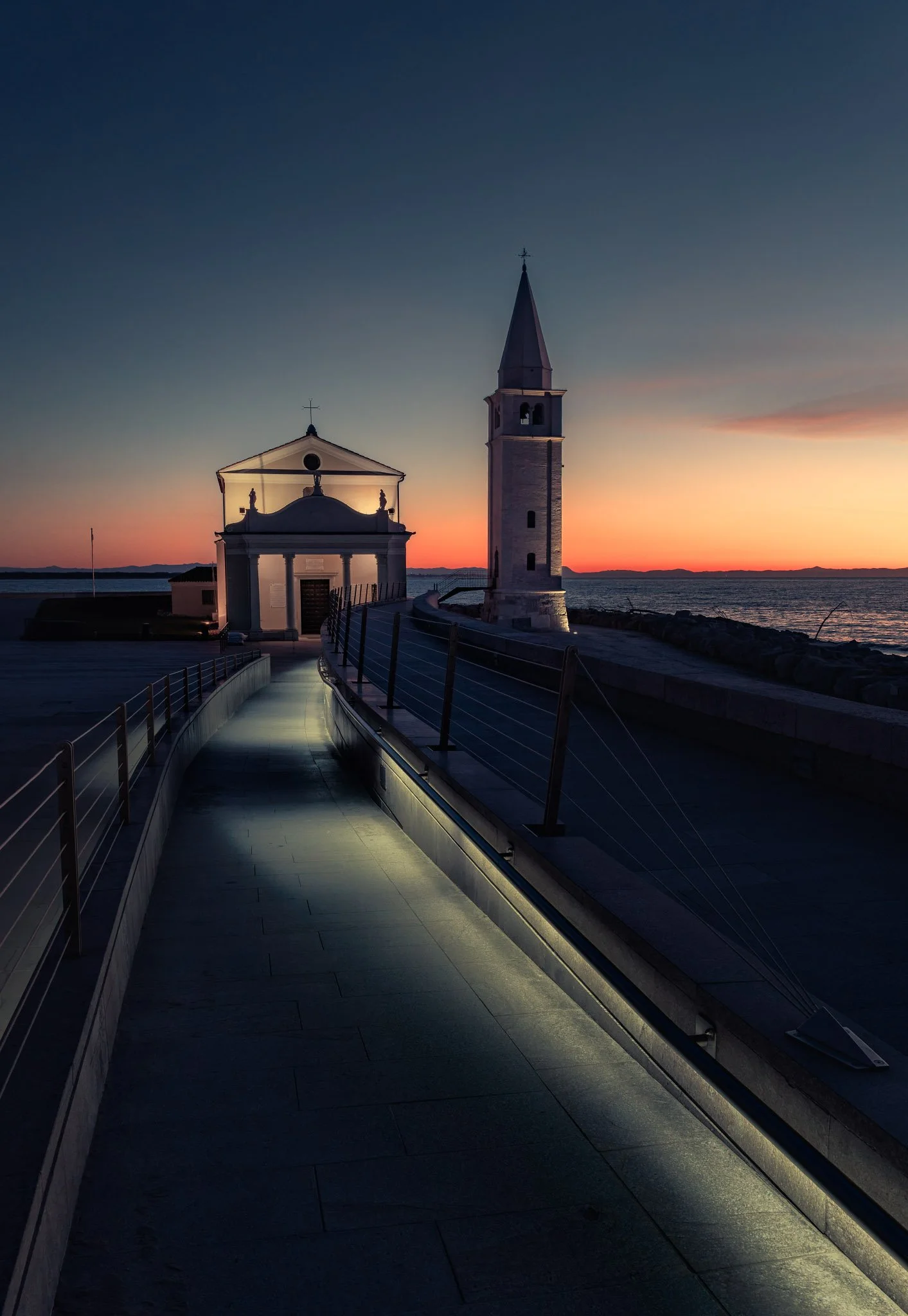 Strada pedonale lungo il mare al tramonto, con torre campanaria e chiesa in stile classico