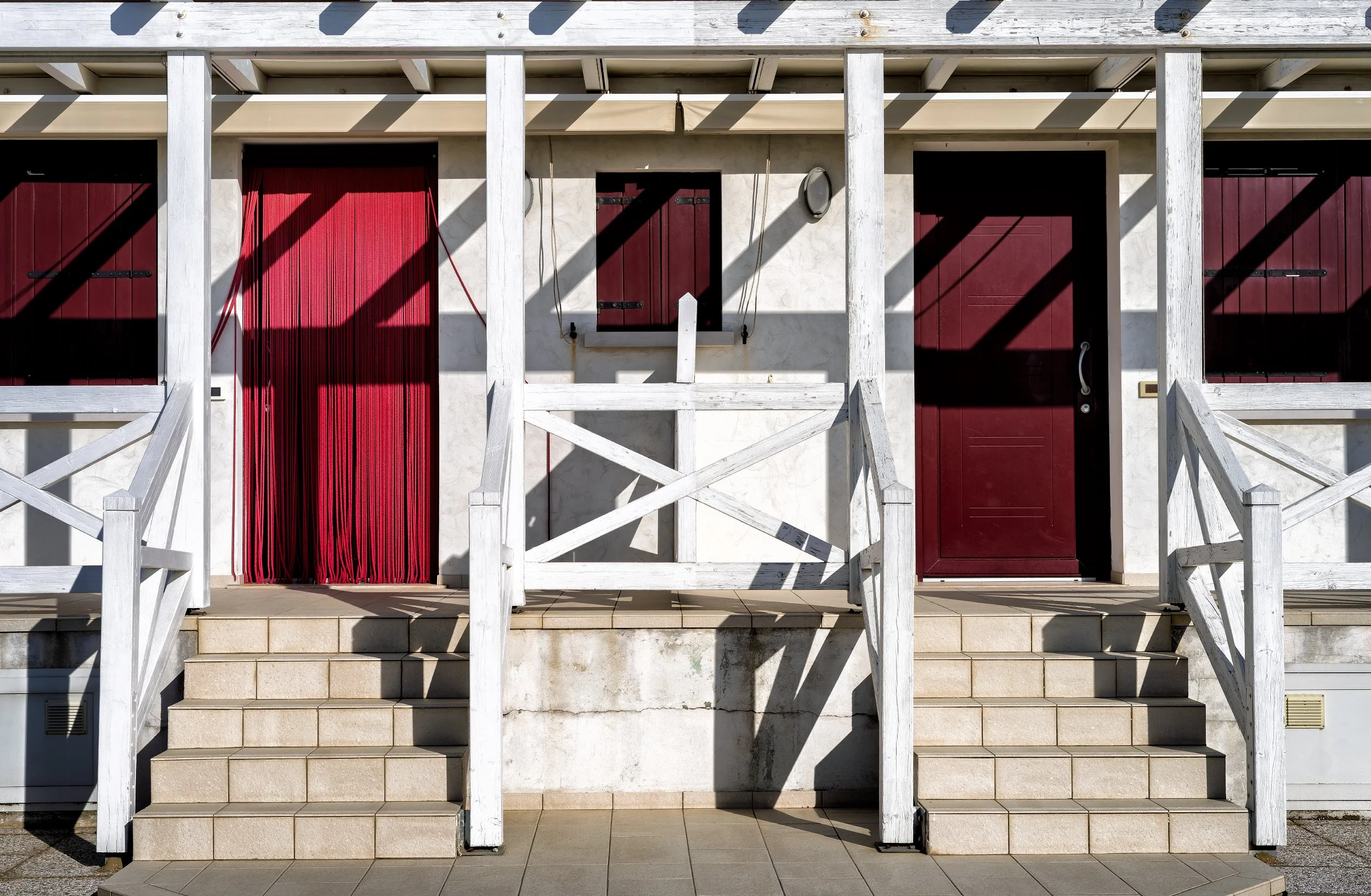 Facciata di un edificio con due ingressi, uno con portone in legno rosso e uno con finestra protetta da persiana rossa, decorata con ringhiere banchina in legno bianco e scale.