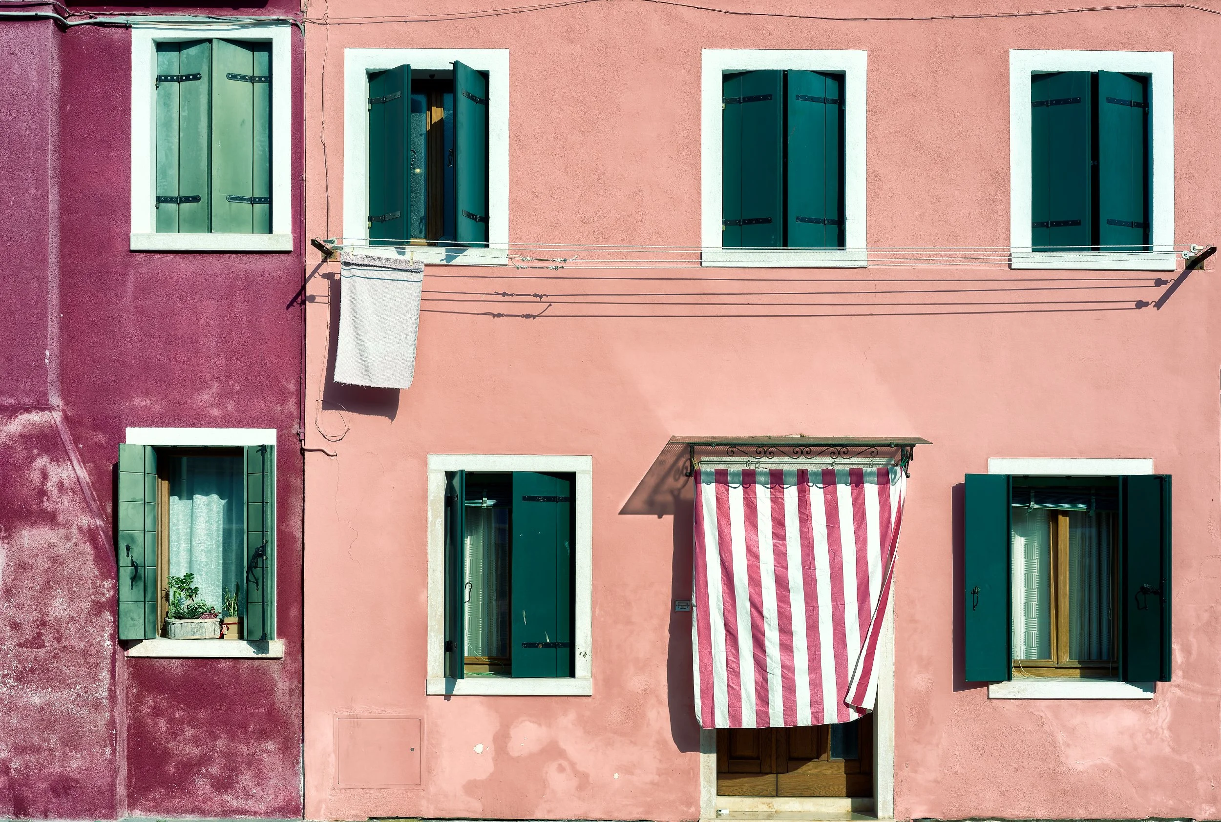 Facciata di un edificio con pareti rosa e viola, quattro finestre con persiane verdi e due balconi, uno con tende a righe rosse e bianche.
