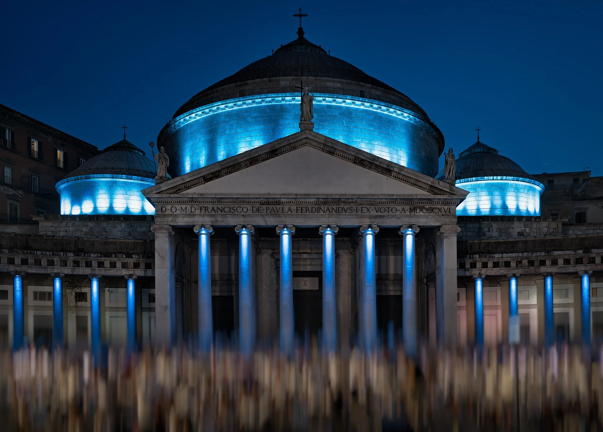 Basilica di San Francesco di Paola illuminata di blu nella notte, con una folla sfocata in Piazza del Plebiscito a Napoli