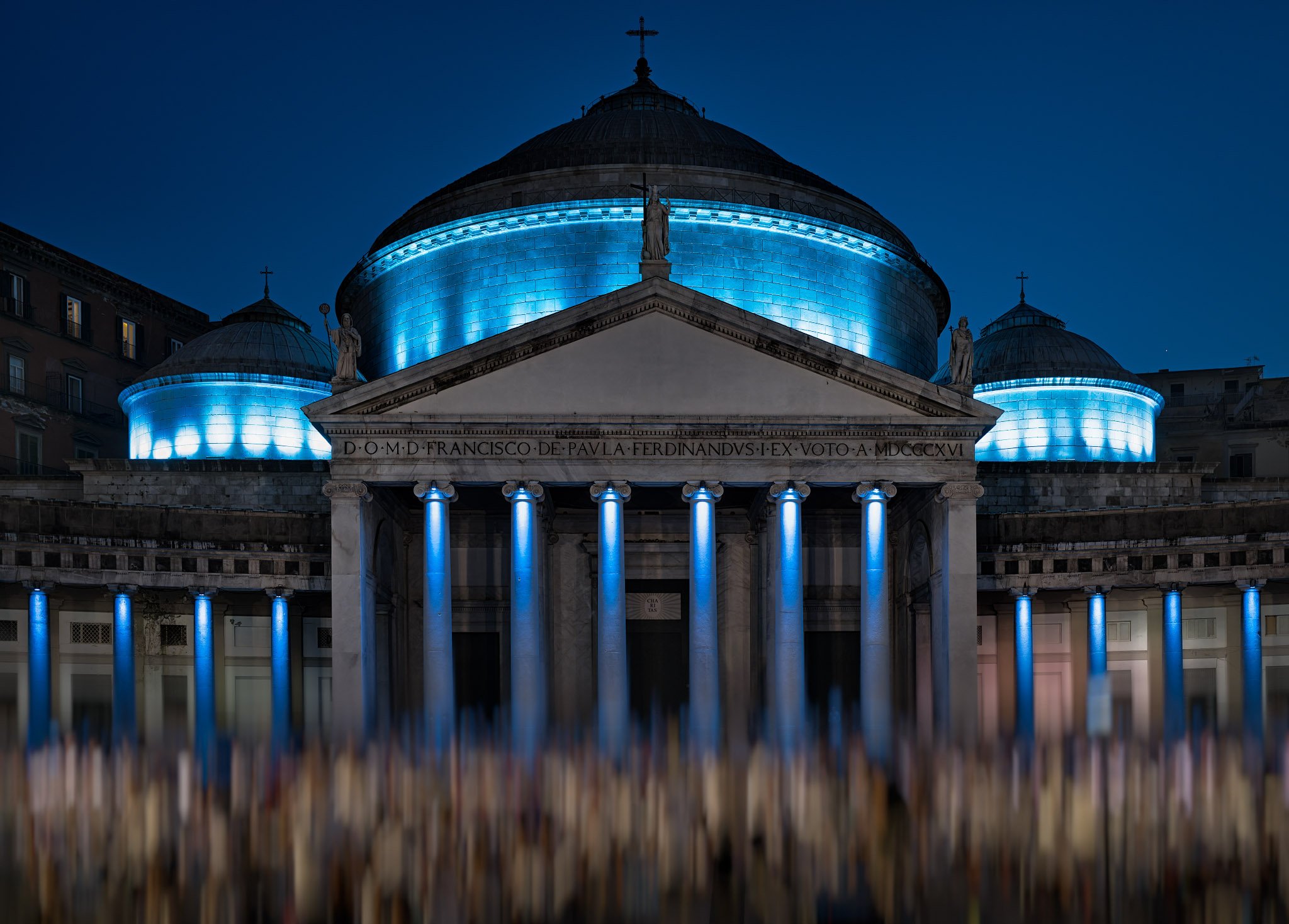 A large audience gathers in front of a monumental building (Basilica of San Francesco di Paola in Naples) illuminated in blue, with columns, a domed roof and a central entrance.