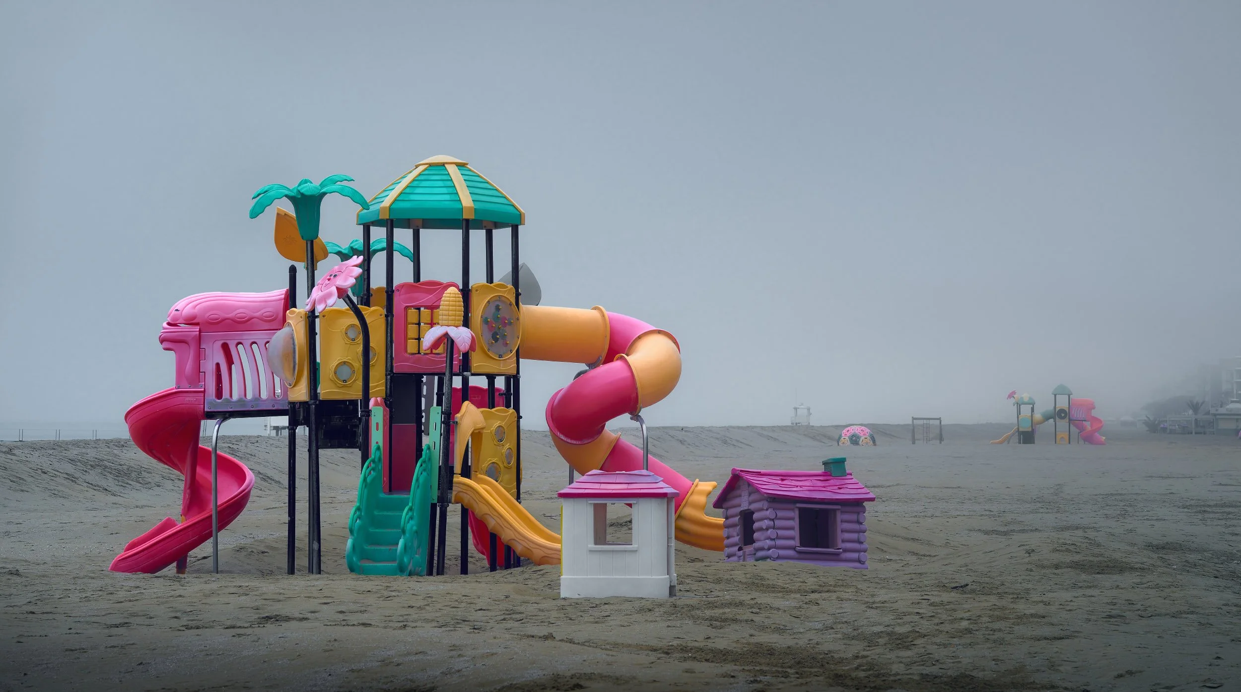 Parco giochi deserto sulla spiaggia con scivoli colorati rosa, gialli, verdi e arancioni, e case di plastica in miniatura, con nebbia e cielo grigio sul mare in lontananza.