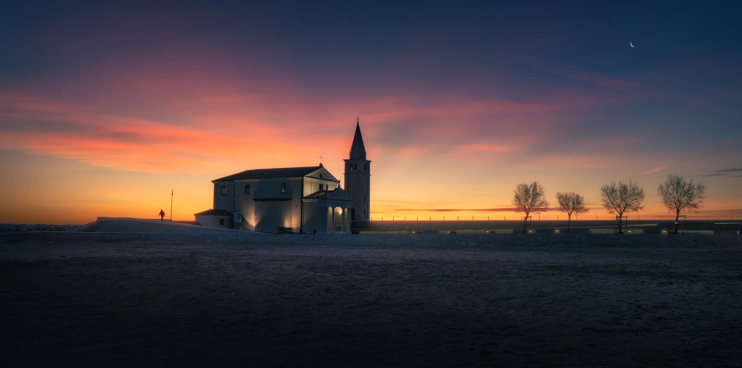 Chiesa con campanile, al tramonto, con quattro alberi spogli sullo sfondo, cielo con sfumature rosa e aranciate, e luna crescente visibile in alto a destra. Una piccola silhouette umana cambia il senso dello spazio attorno alla chiesa.