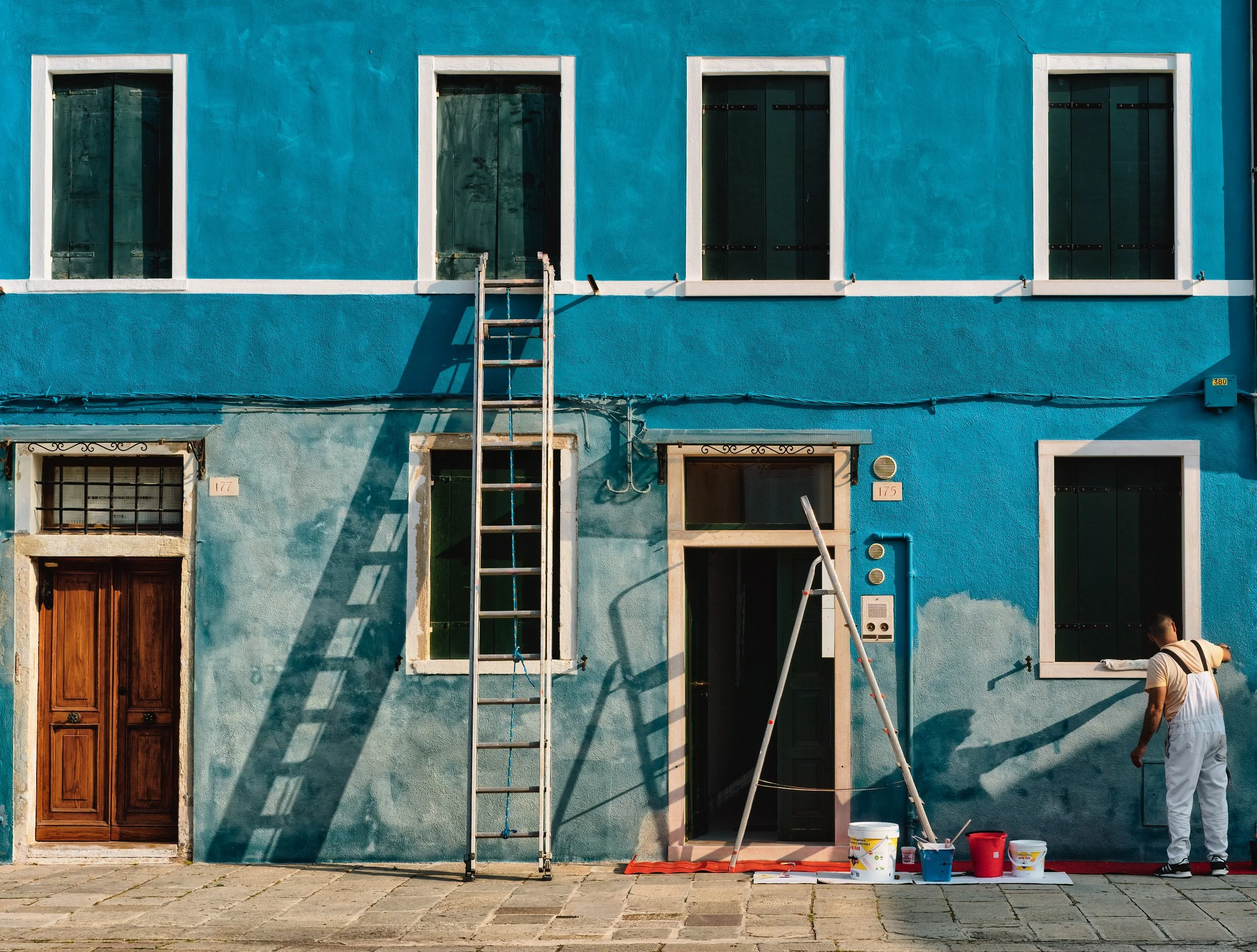 Veduta di una facciata di edificio con quattro finestre e una porta, in fase di pittura, con un uomo che lavora alla parete e attrezzi per pittura sul pavimento.