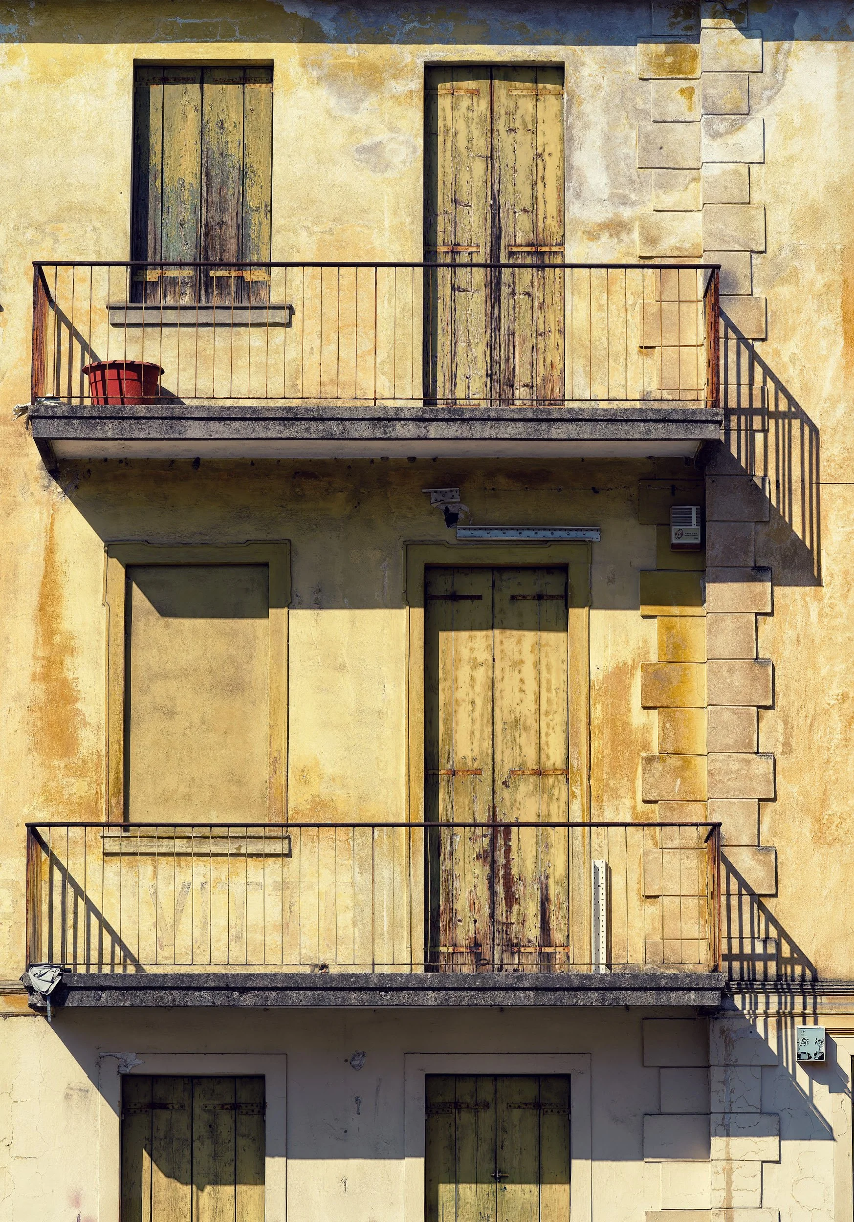 Facciata di un edificio con quattro balconi sovrapposti, con pareti gialle e persiane in legno chiuse, alcune delle quali rovinate o scolorite. Una delle aperture risulta murata.