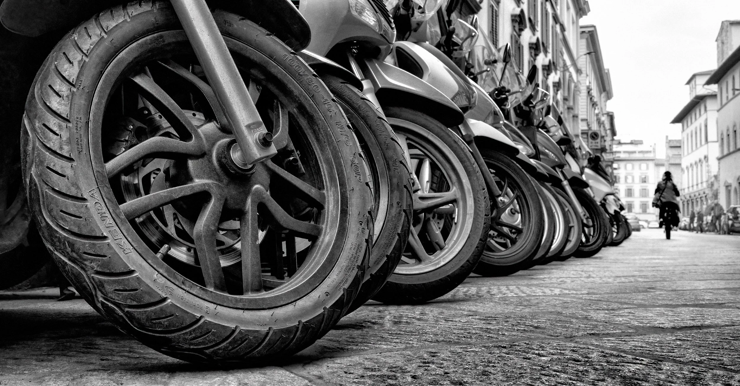 A person rides a scooter past a row of parked bicycles and scooters, with overlapping wheels and metal frames creating a layered urban pattern.