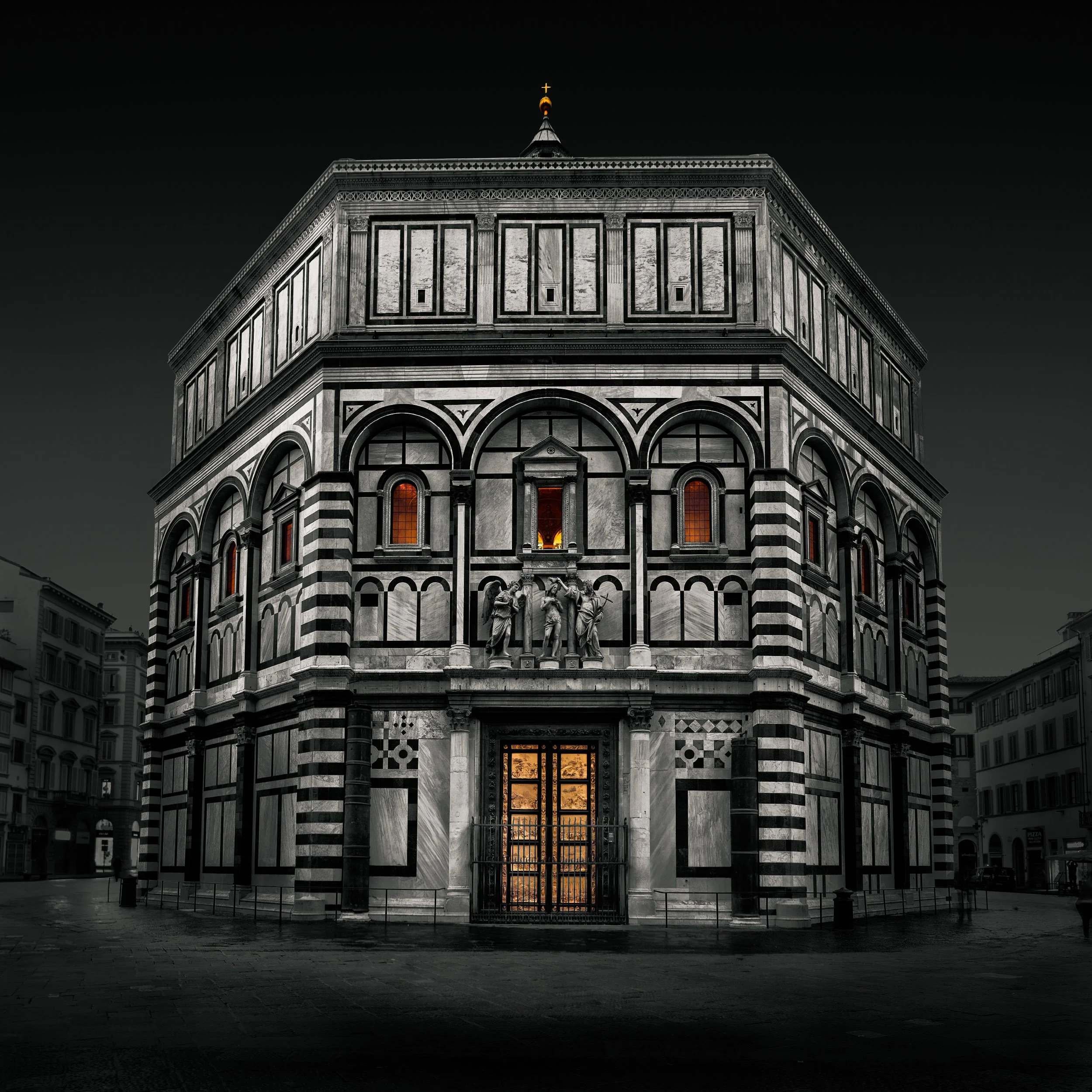 Una chiesa in pietra bianca (il Battistero di Firenze) con dettagli architettonici eleganti, alcune statue sulla facciata e finestre con vetri arancioni illuminati, vista notturna.