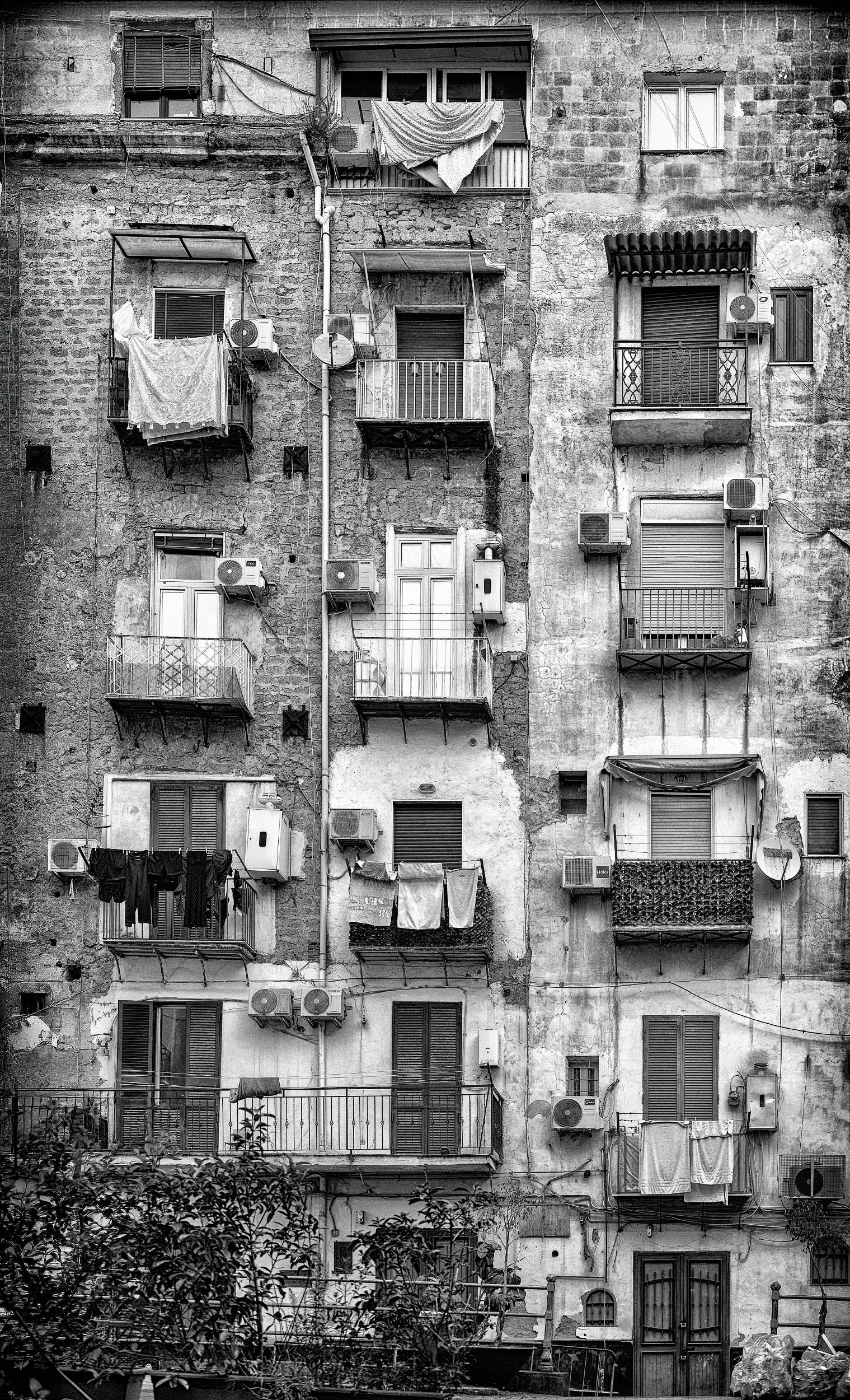 Facciata di un edificio antico con molte finestre, balconi e aria condizionata esterna. Pareti in pietra e calce, alcune con bucature e segni di usura. Tende, lavatrici e vari oggetti sui balconi.