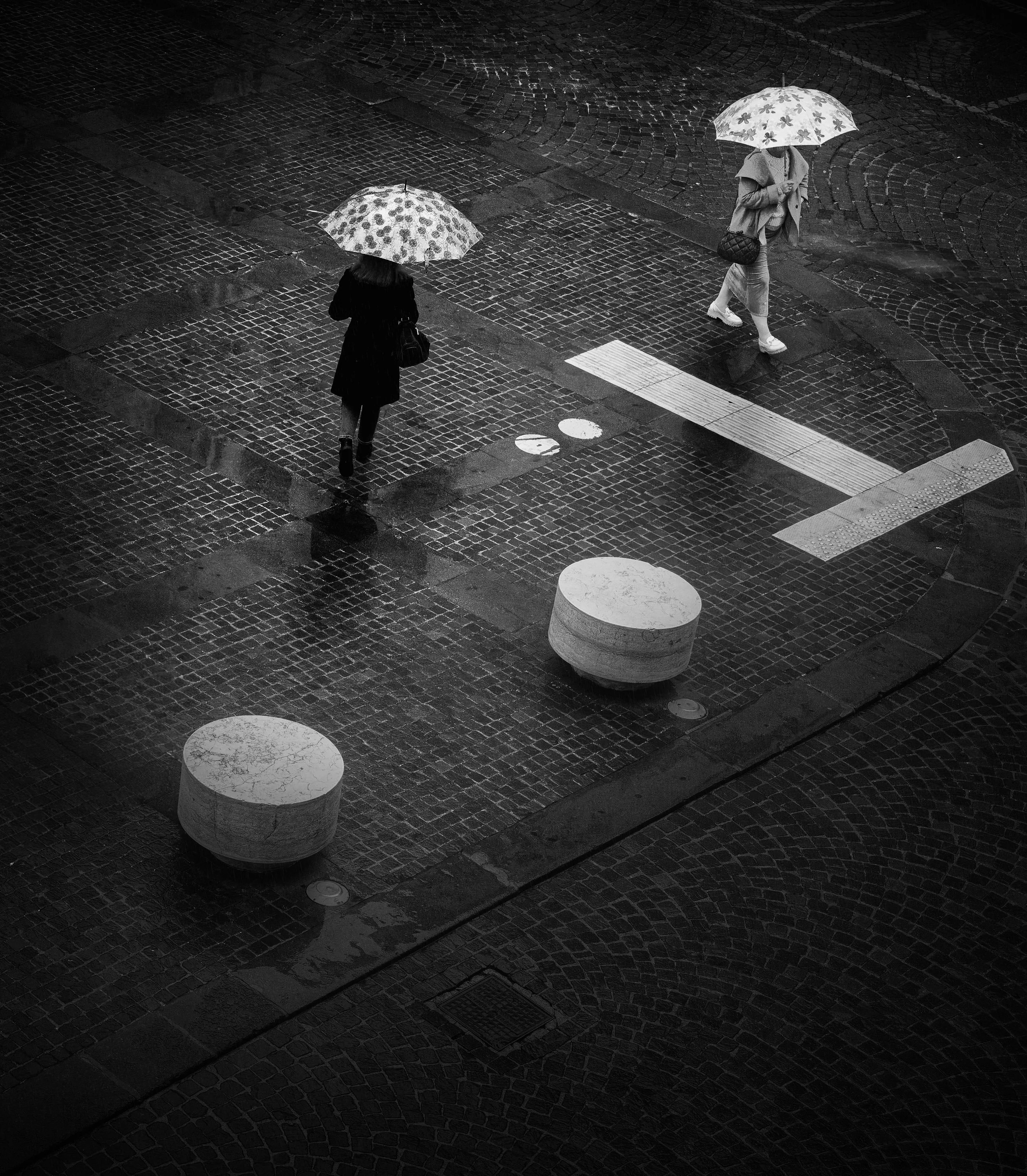 Due persone camminano sotto un ombrello sulla strada bagnata di notte, vista dall'alto in bianco e nero. Gli ombrelli richiamano forma e geometria di due divisori stradali.