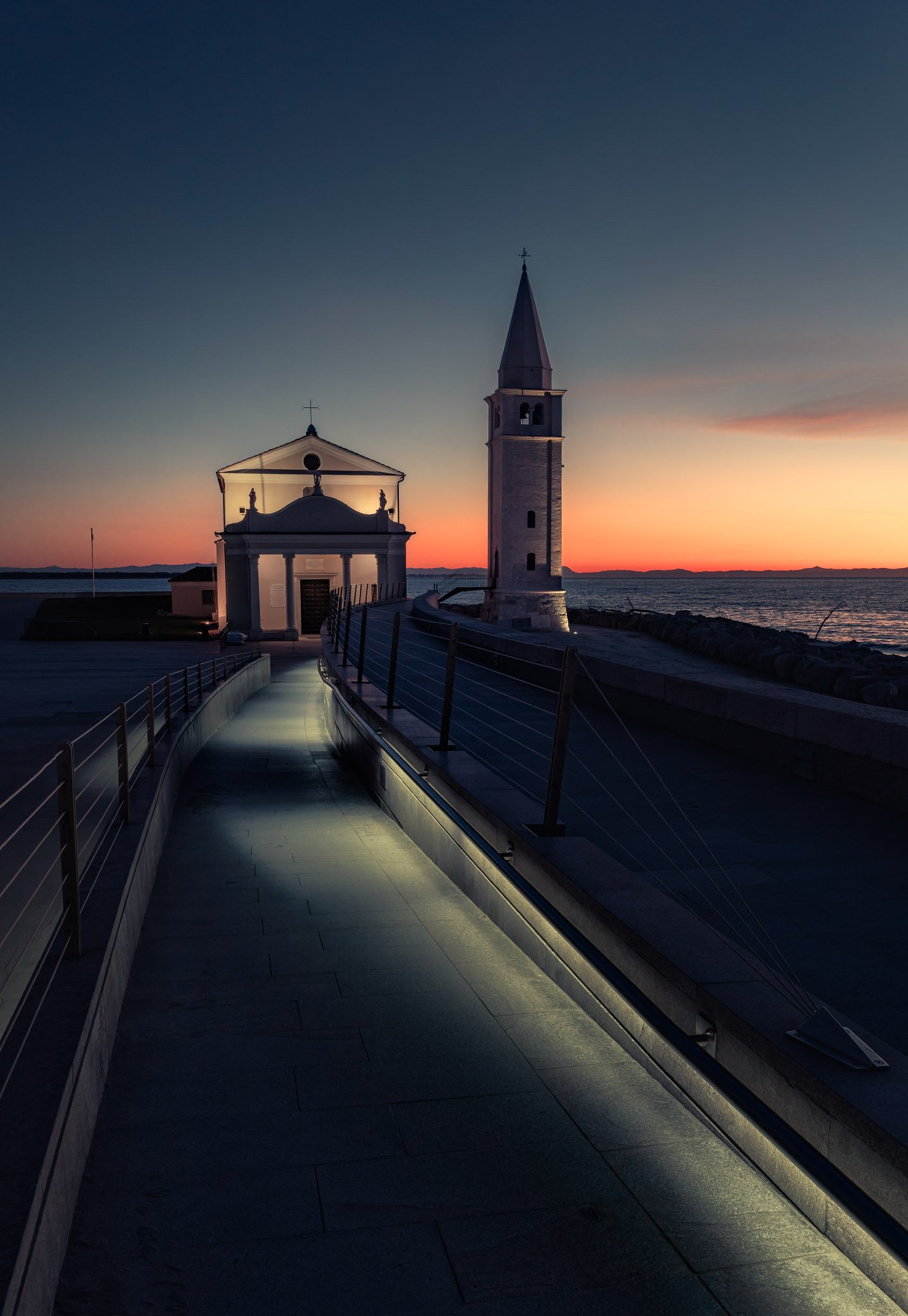 A illuminated walkaway leads towards a small white chapel and bell tower beneath a blue and orange dawn sky.