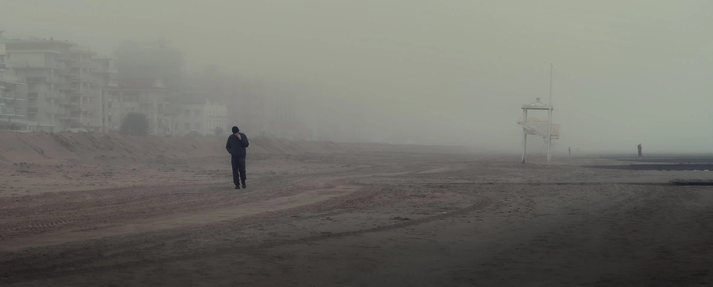 Uomo che cammina sulla spiaggia deserta sotto una nebbia densa, con alcune persone distanti e un'area con una torretta di salvataggio.