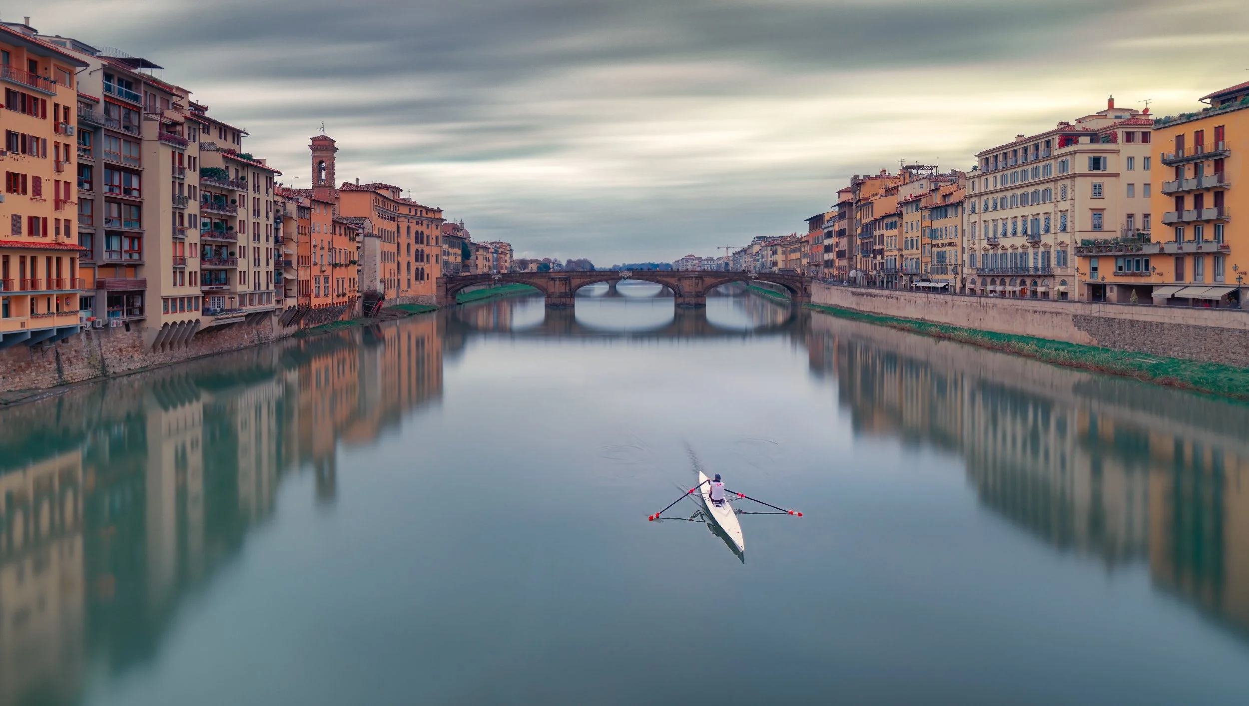 Una scena serale di un fiume con edifici colorati lungo le sue sponde e un ponte in pietra al centro. Nel fiume, una persona pratica il paddleboarding, con l'acqua che riflette le costruzioni e il cielo nuvoloso.