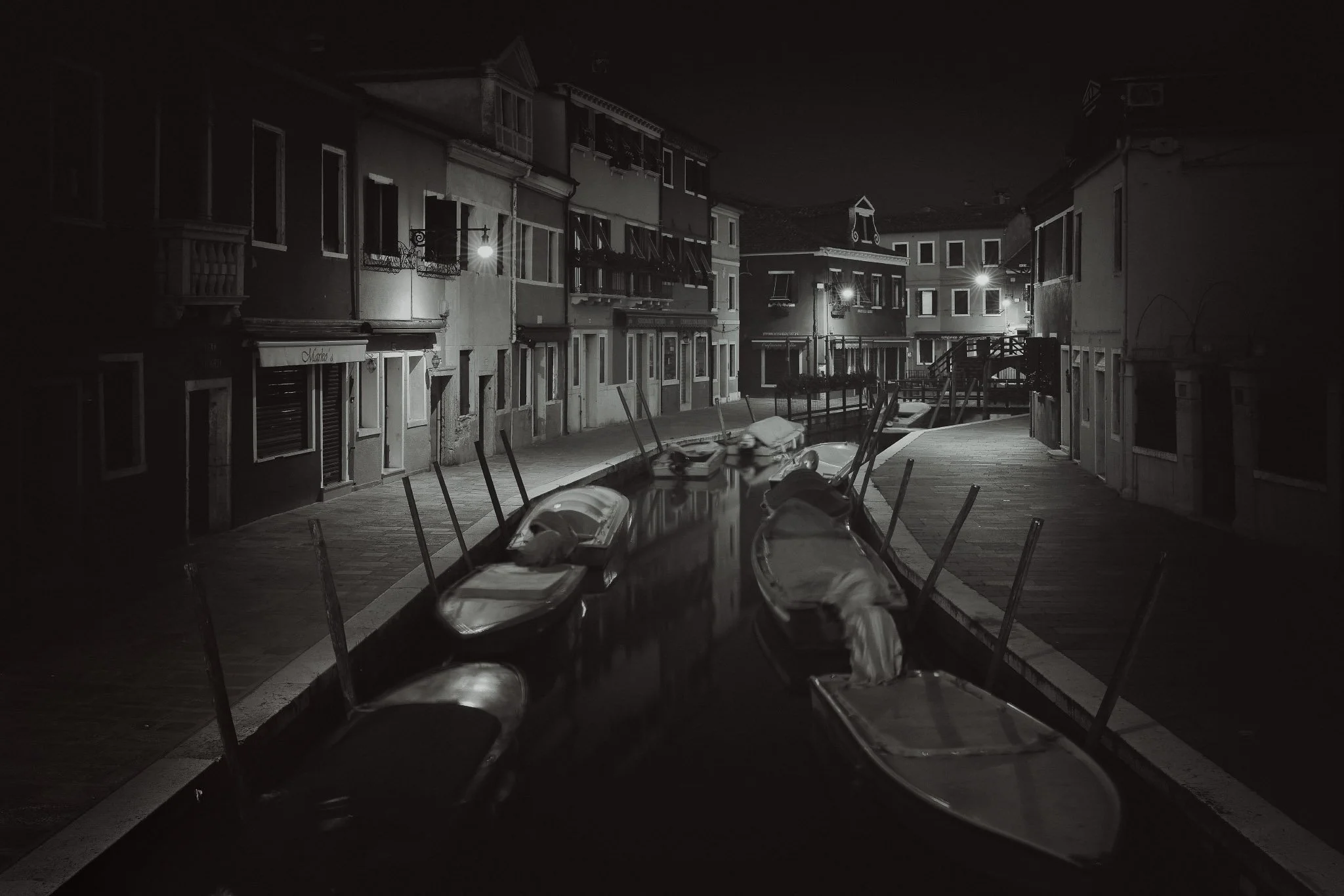 A quiet Burano canal at night in black and white, lined with houses, moored boats and war street lights reflected in the dark water.