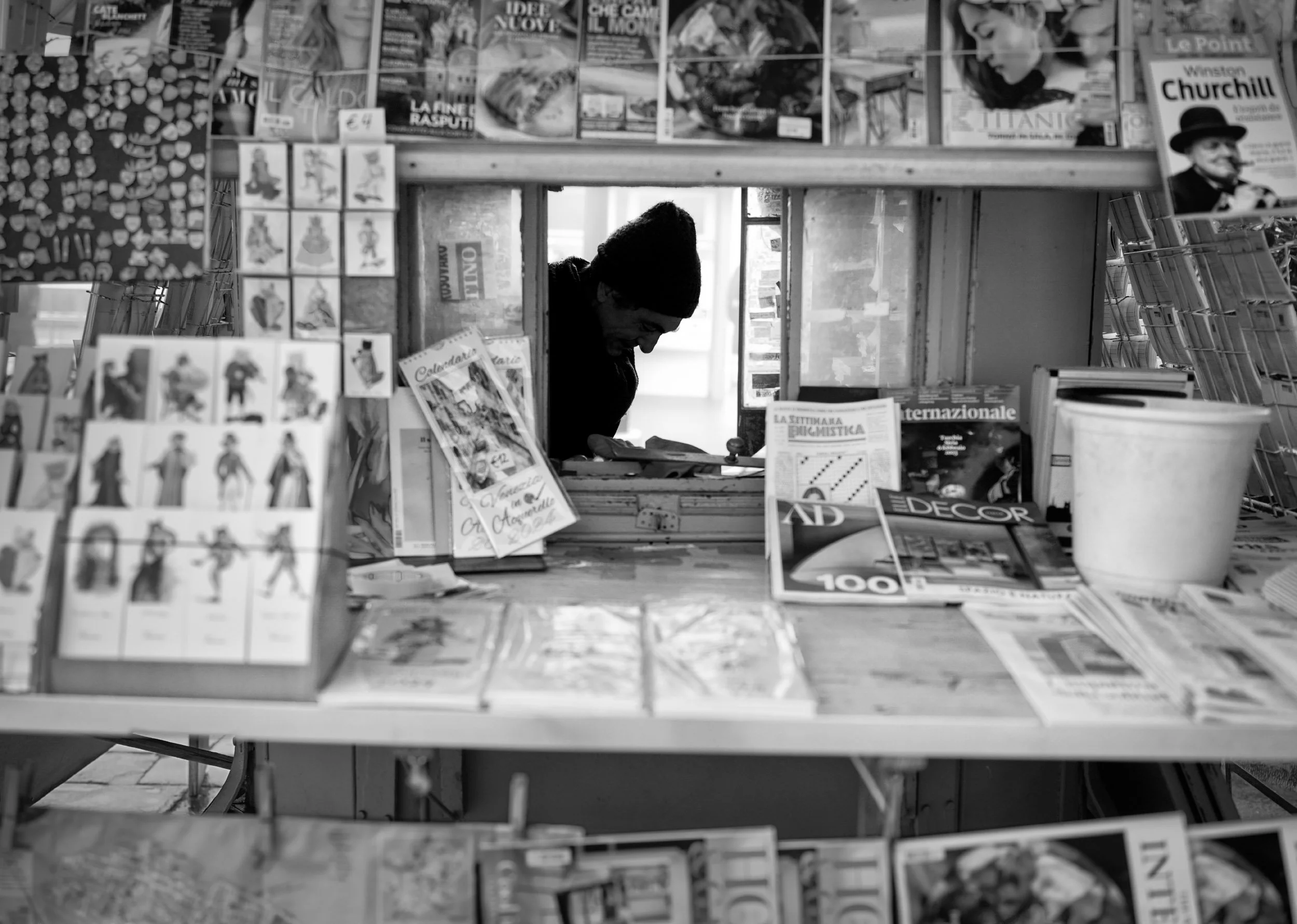 A newsagent sits inside a kiosk, surrounded by newspaper, magazines, postcards and printed images.