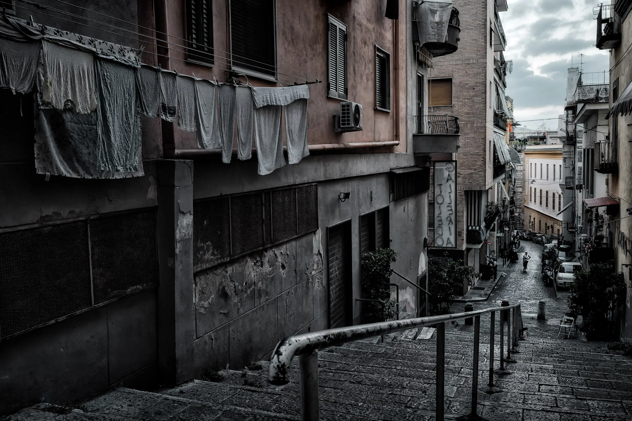 Vista di un vicolo cittadino con edifici vecchi e ammaccati, alcune balconate, biancheria stesa ad asciugare e un cartellone con scritte. Cielo nuvoloso, strada in pendenza con alcune persone e veicoli parcheggiati.