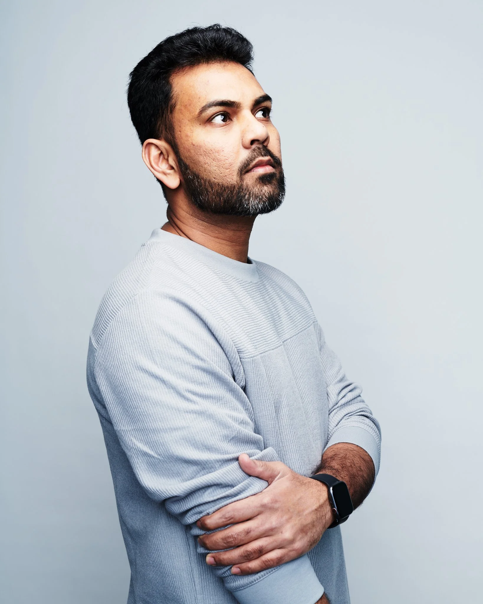 Praveen Sake looks to his right with crossed arms, wearing a light gray sweater and a smartwatch on his left wrist, against a plain light gray background.