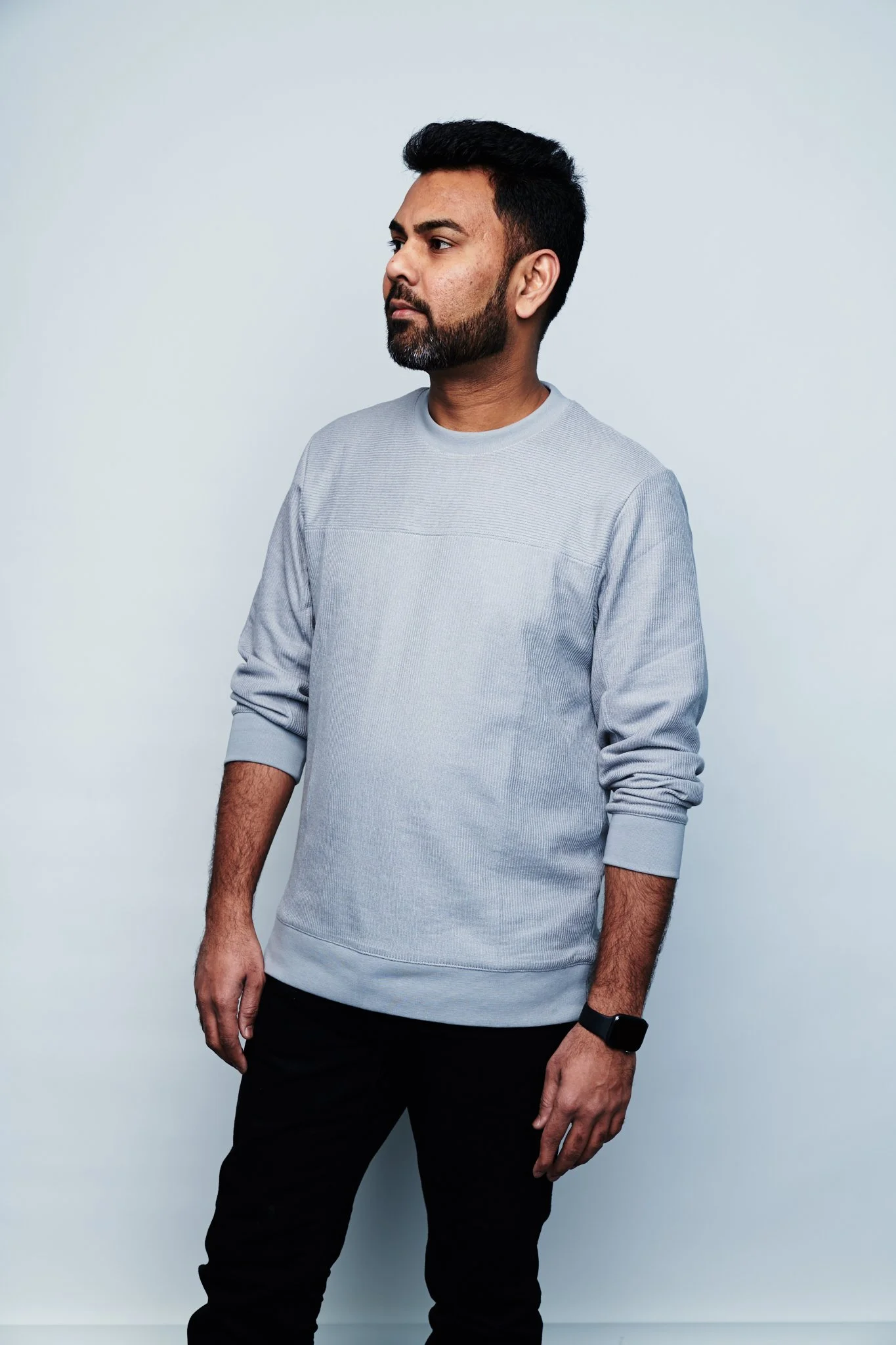 Praveen Sake standing against a plain light blue background, wearing a light gray long-sleeve shirt and black pants, with a black smartwatch on his left wrist.
