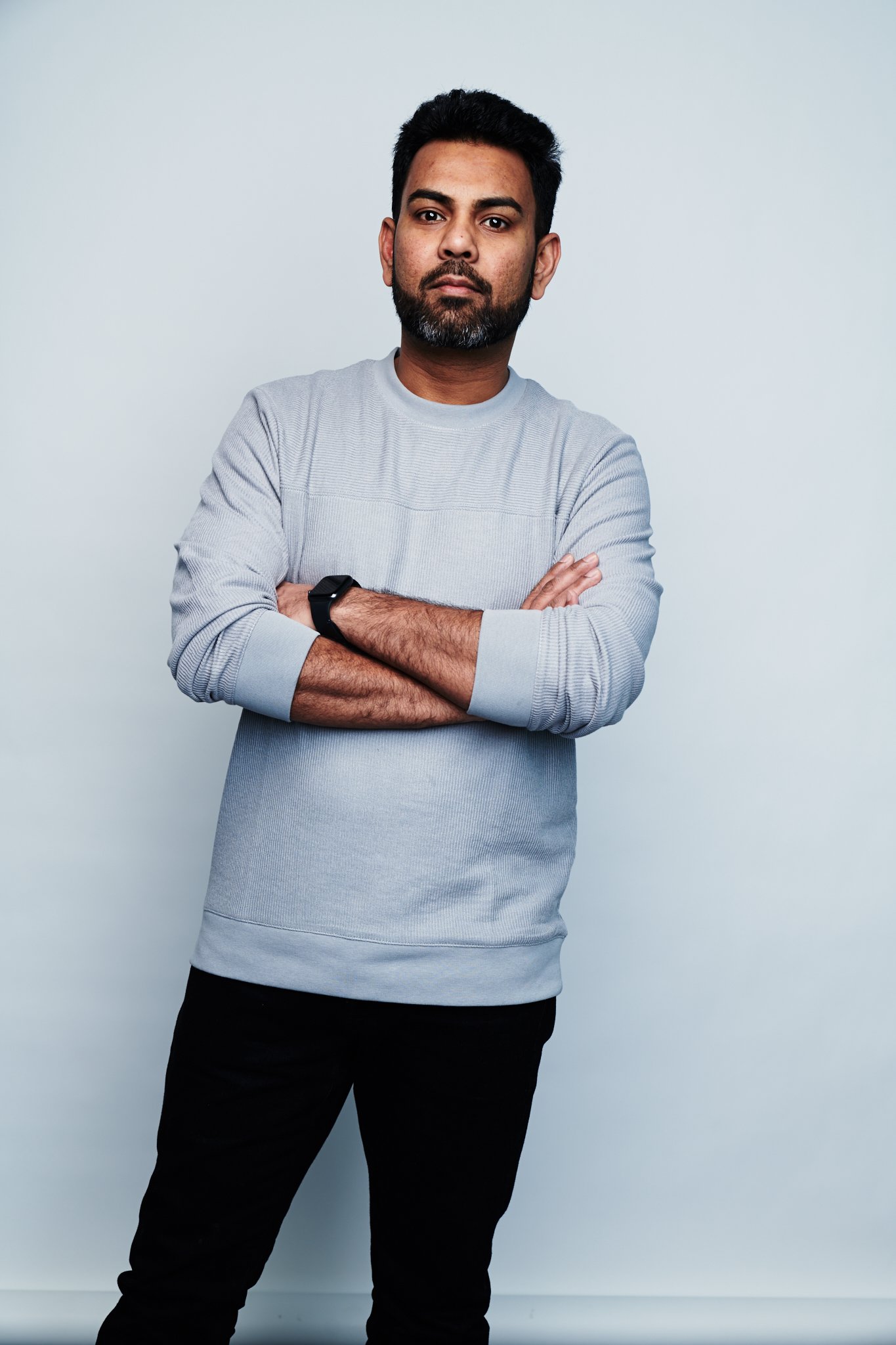 Praveen Sake with crossed arms wearing a light gray sweater and black pants, standing against a plain background.